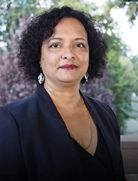 A woman with curly black hair, wearing a dark blazer and earrings, standing outdoors with greenery in the background.