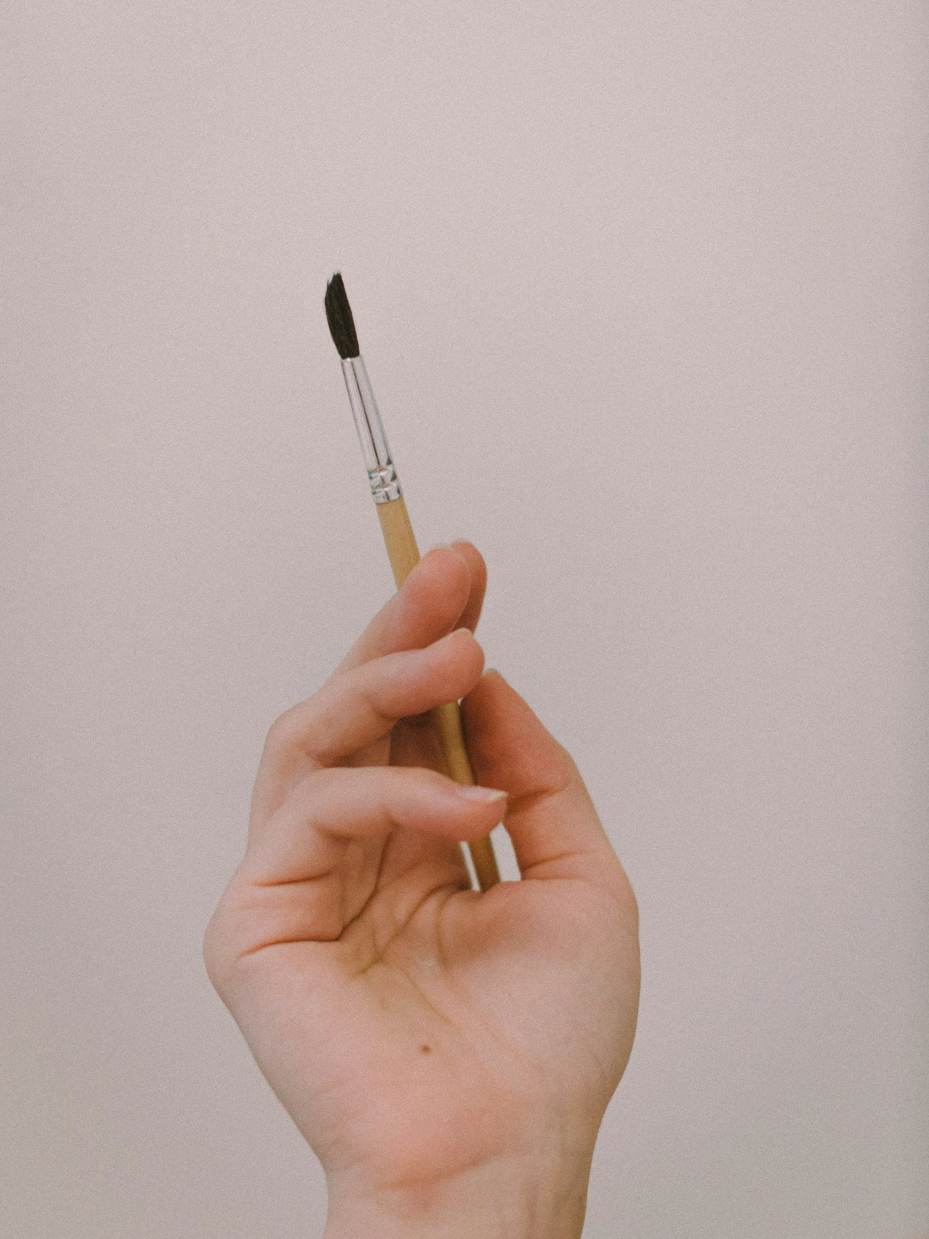 A hand holding a paintbrush with black bristles against a plain, light-colored background.