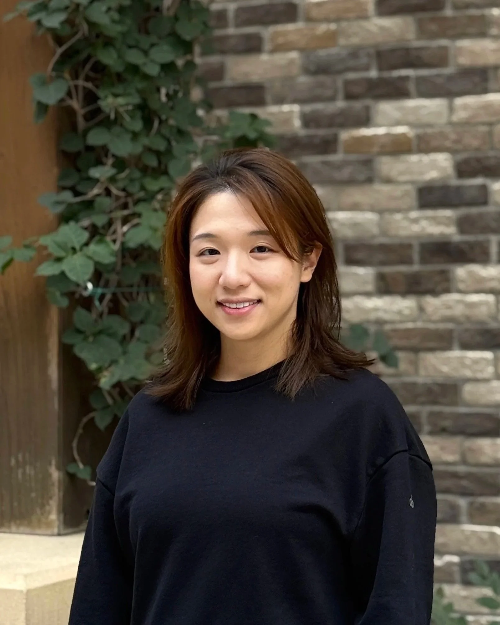 A woman with shoulder-length brown hair, wearing a black shirt, standing outdoors in front of a brick wall and leafy green plants, smiling at the camera.