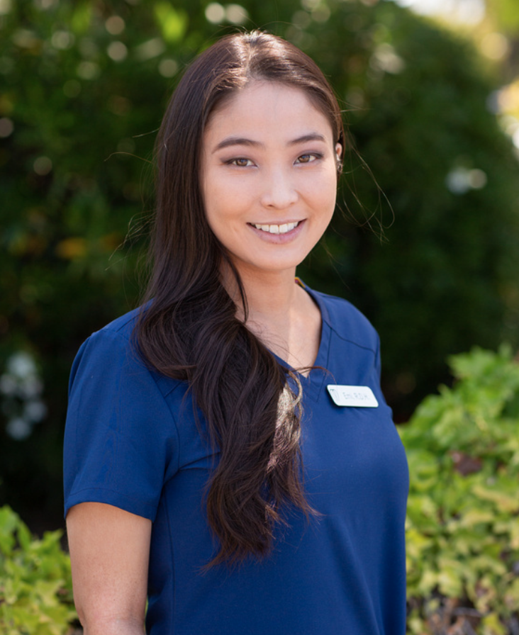 A young woman with long dark hair wearing a blue uniform and name tag, standing outdoors surrounded by greenery, smiling at the camera.