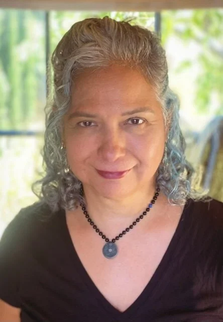 A woman with gray, curly hair and light skin, wearing a black top and a beaded necklace, smiling indoors with a window showing green foliage in the background.