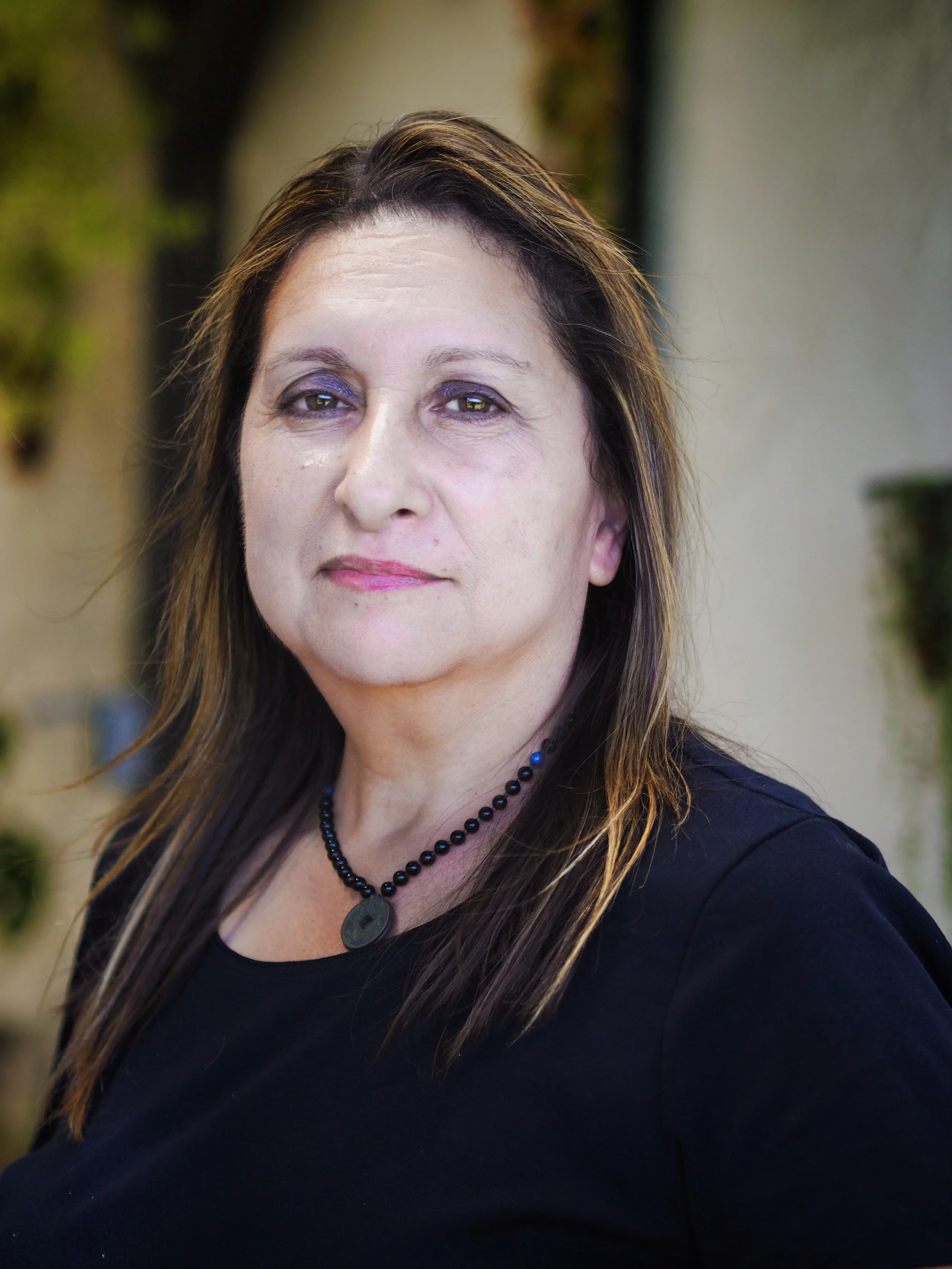 A woman with long brown hair and purple eye makeup, wearing a black top and a black beaded necklace, standing outdoors with blurred greenery and buildings in the background.
