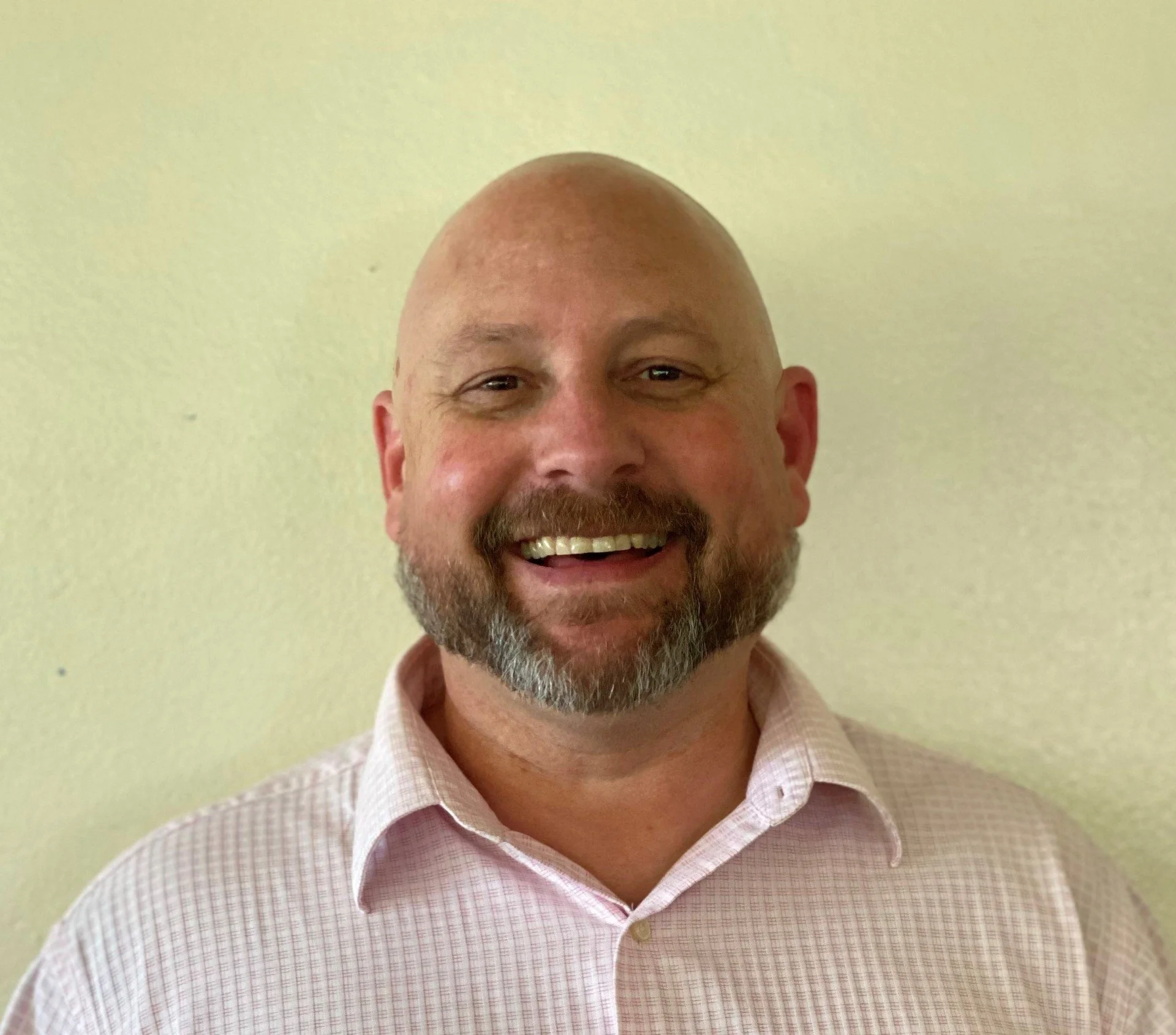 A smiling middle-aged man with a beard and bald head, wearing a light pink checkered shirt, standing against a plain light-colored wall.