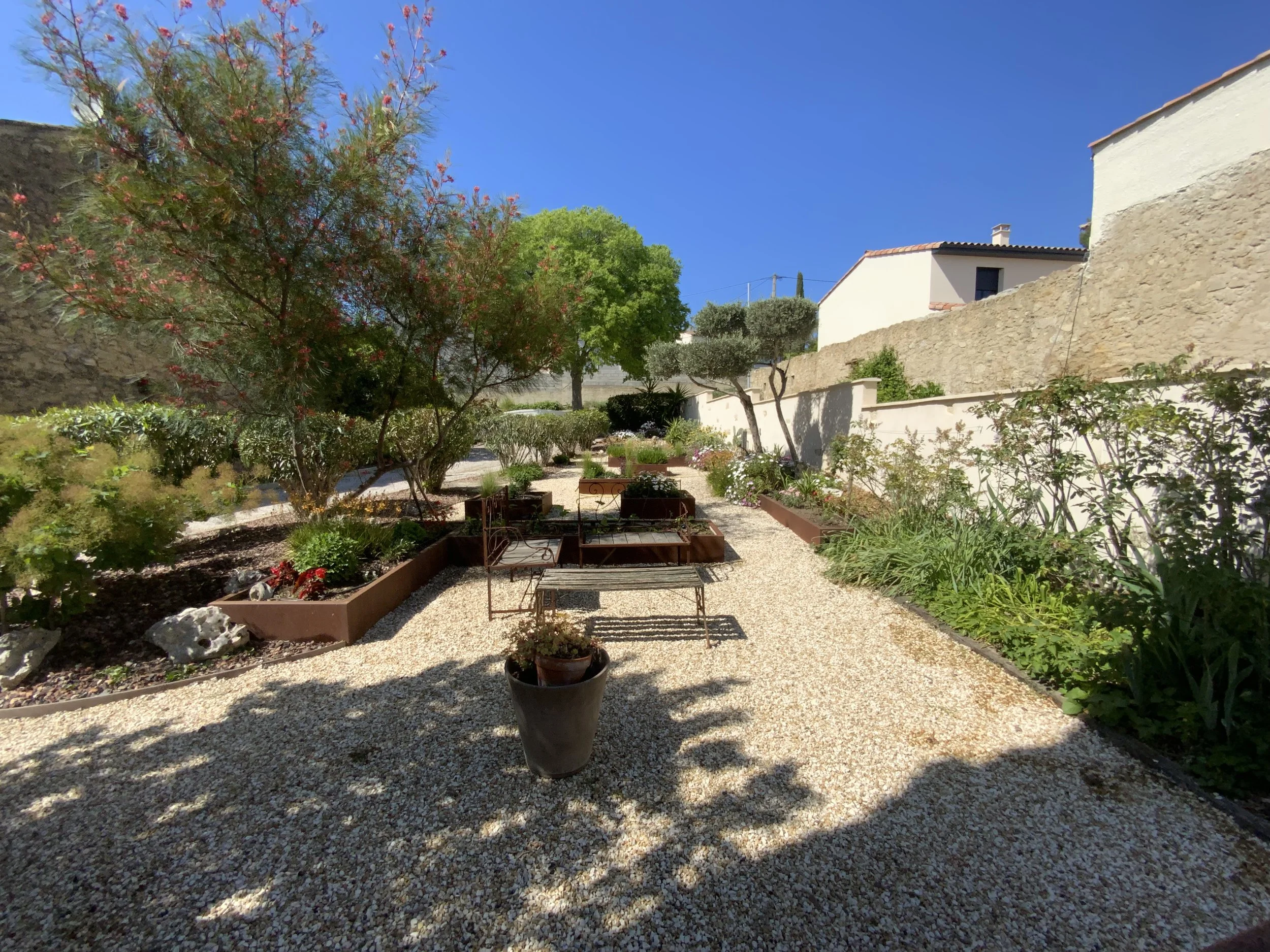 Un jardin avec des arbres, des arbustes et des fleurs, des bancs en métal, un mur en pierre et des maisons blanches avec un toit en tuiles rouges sous un ciel bleu clair.