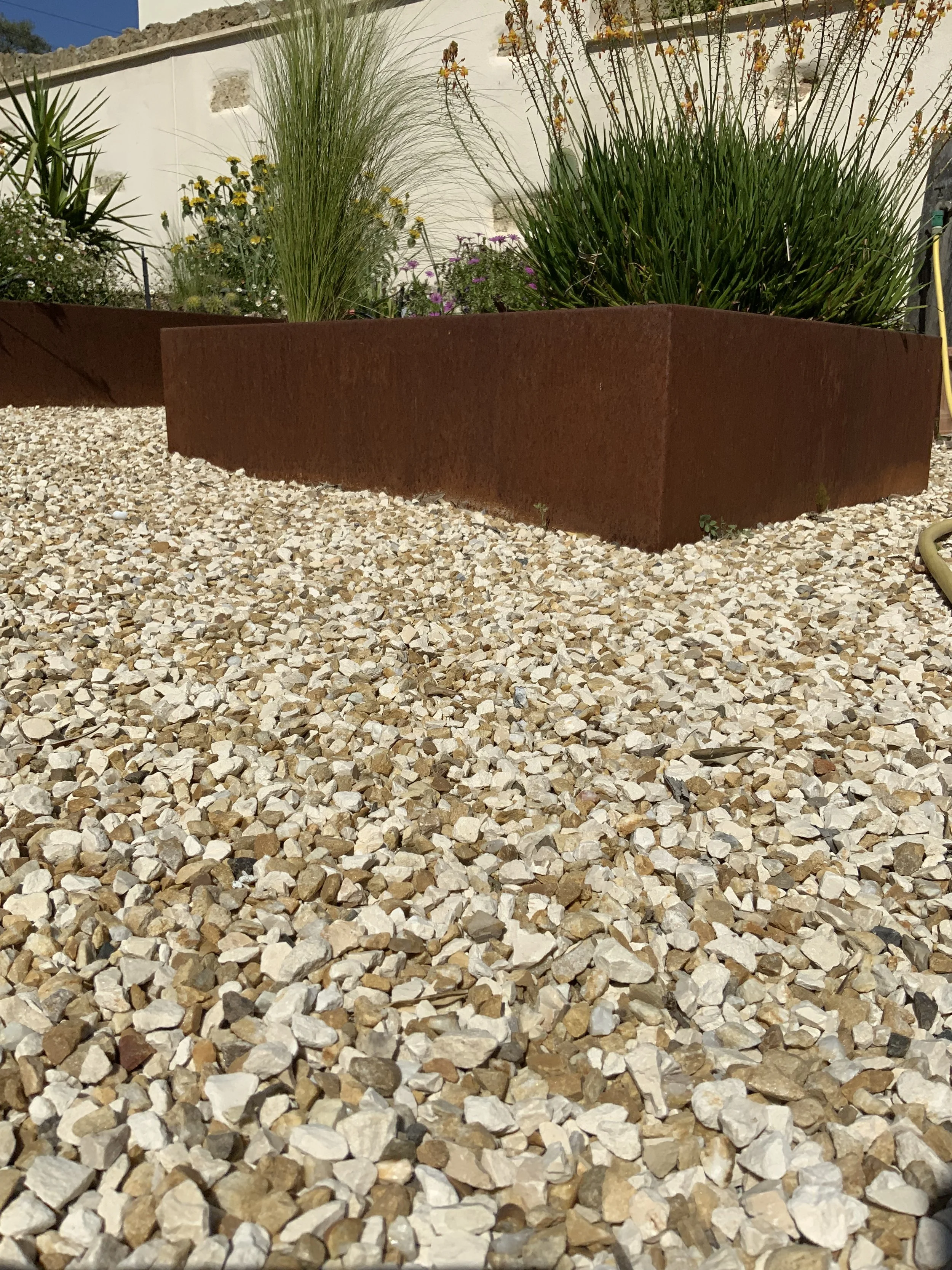 Un jardin avec des plantes dans un bac en métal rouillé posé sur un sol recouvert de petits cailloux, avec un mur blanc en arrière-plan.