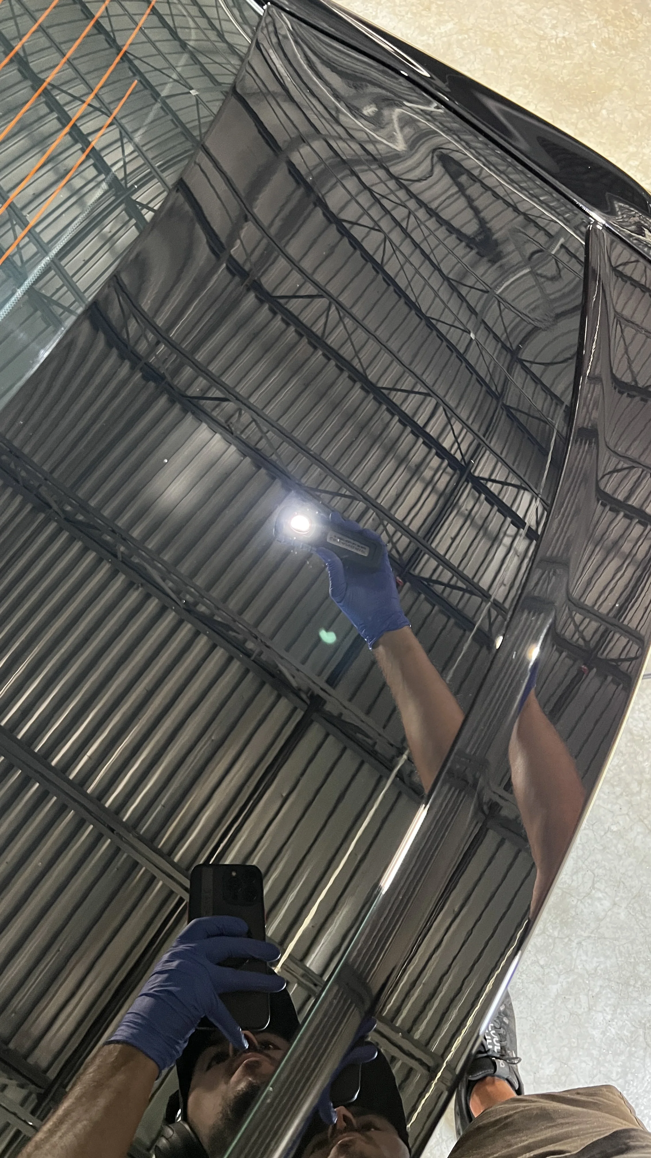 Person taking a photo of a reflective black appliance, possibly a refrigerator, with a smartphone. The person is wearing blue gloves and a black cap, and is in an industrial or warehouse setting.