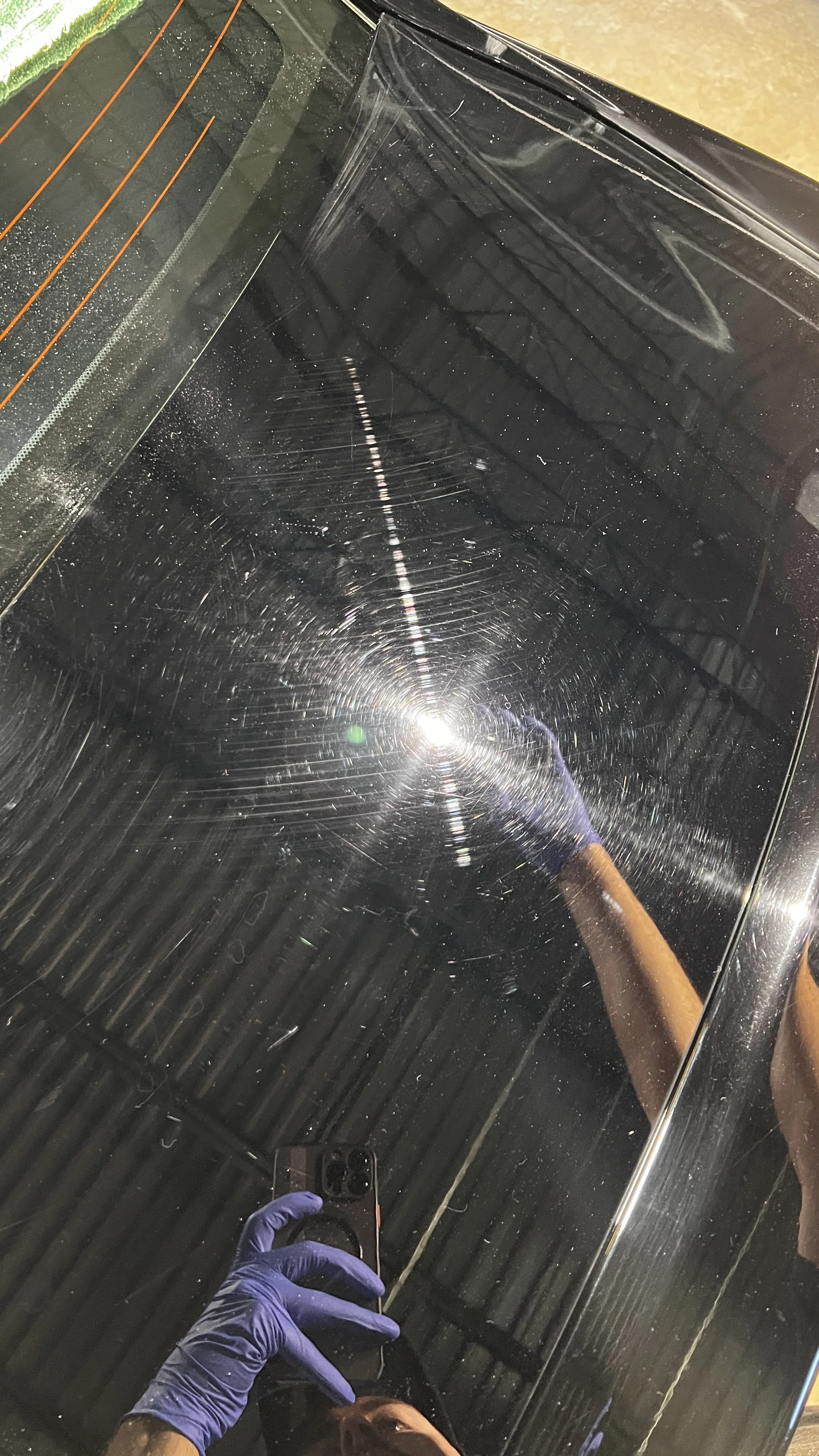 Close-up of a scratched black glass or mirror surface with reflections of the person taking the photo, wearing purple gloves, and factory ceiling lights and structures in the background.