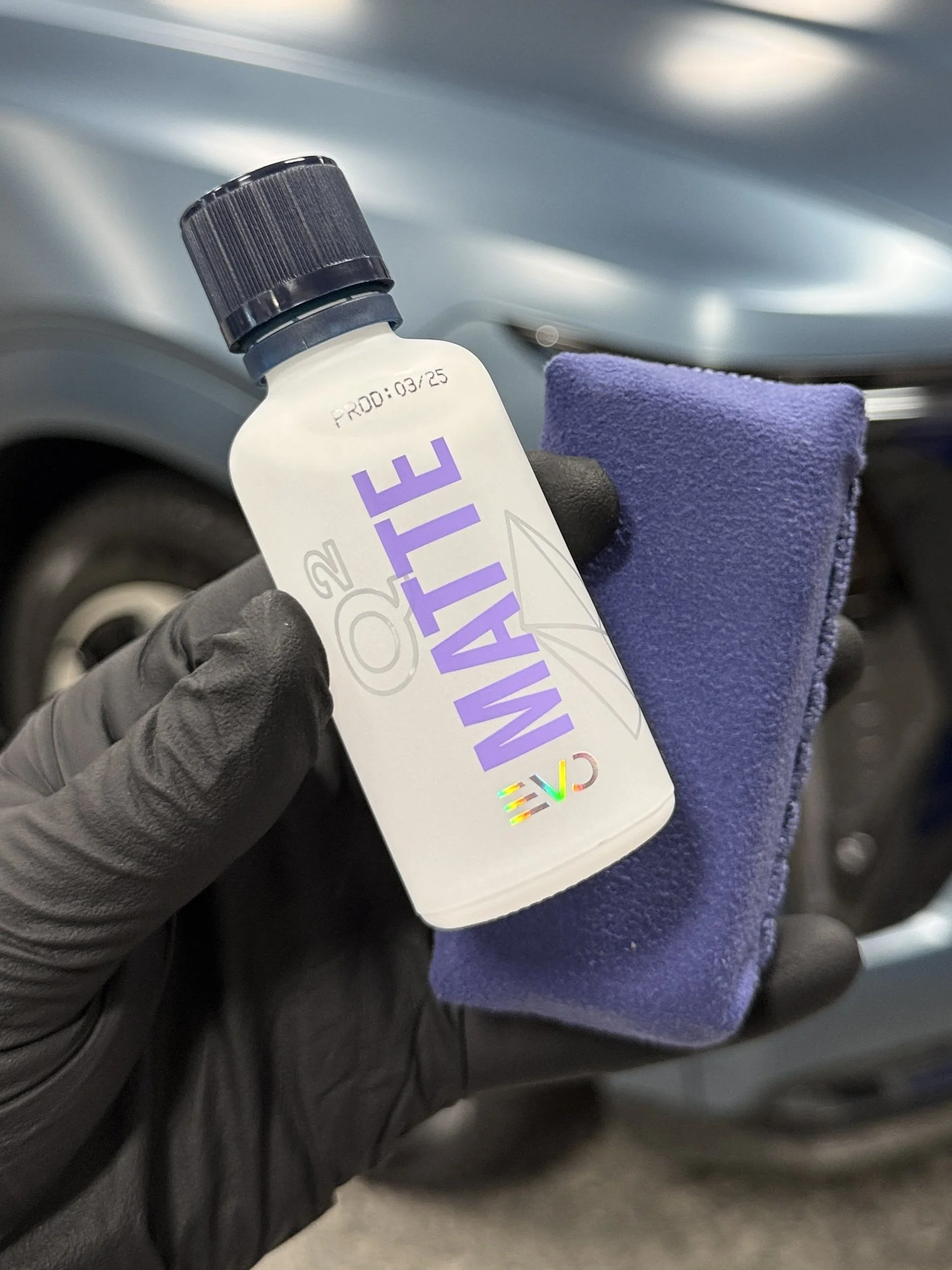 A hand wearing a black glove holding a small spray bottle labeled 'MALIT' with a purple and holographic logo, along with a purple microfiber cloth, in front of a motorcycle wheel.