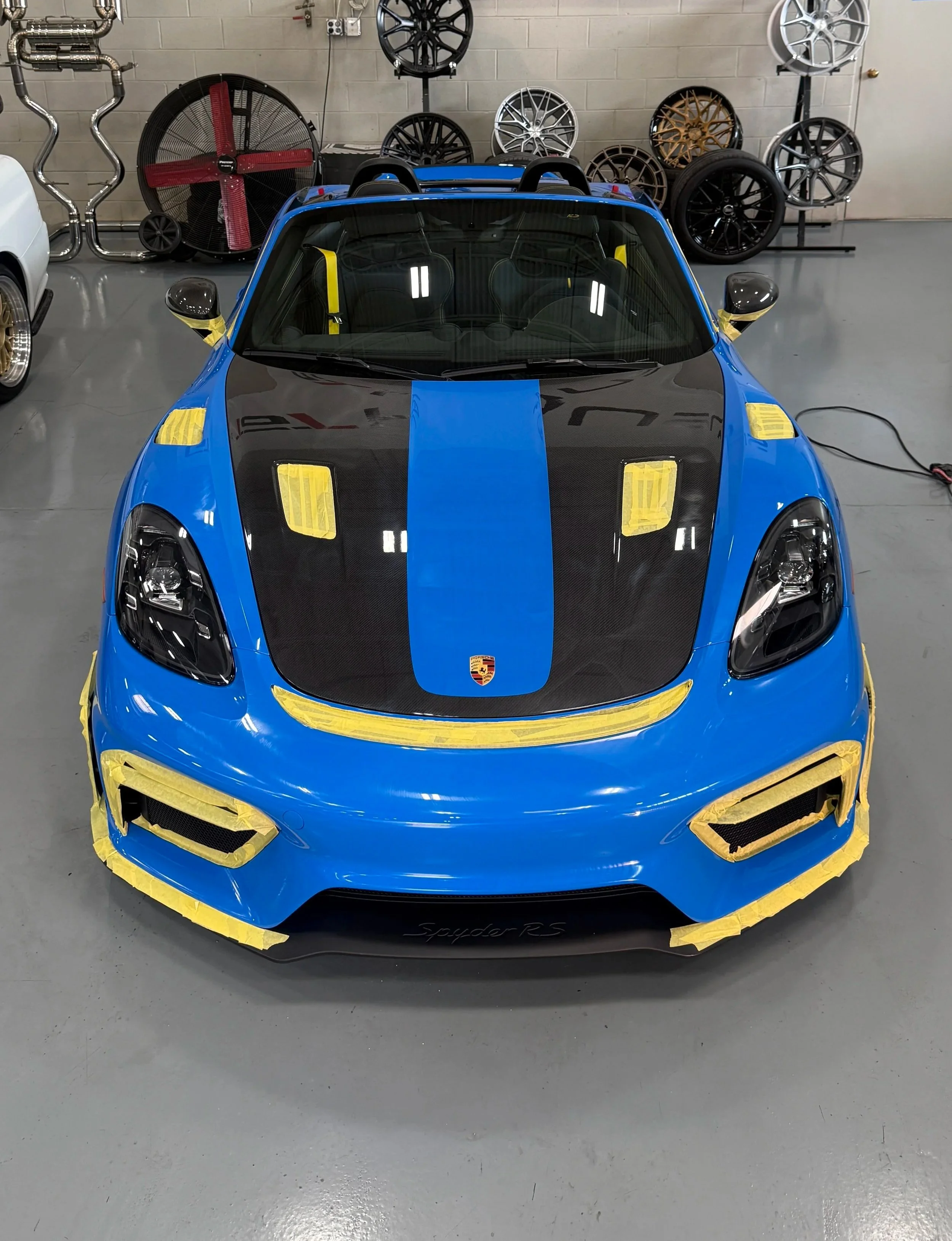 Blue sports car with black and yellow accents in a garage, surrounded by various car rims and wheels on the wall.