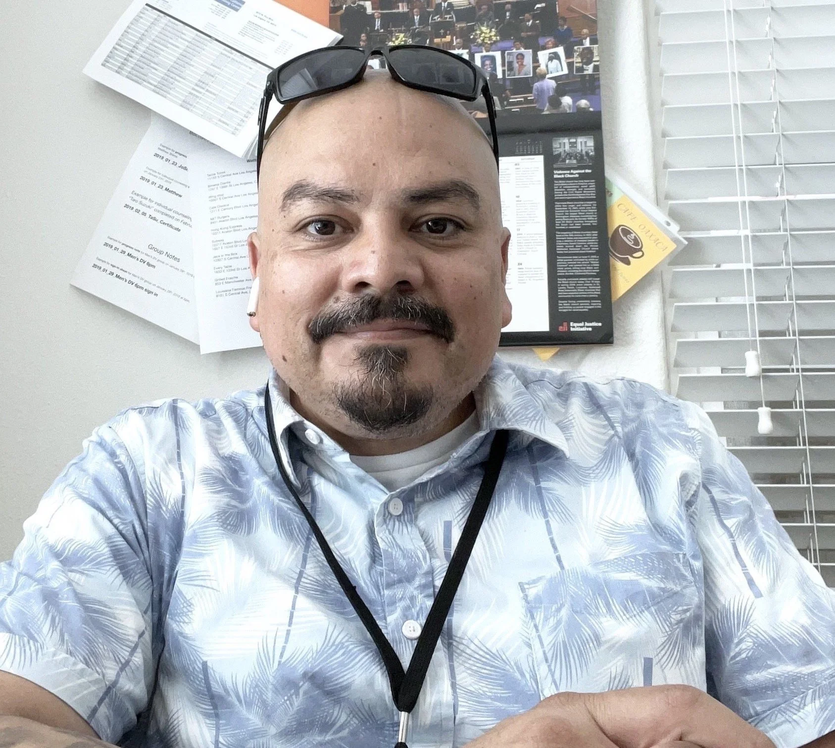 A man taking a selfie in an office, wearing a button-up shirt with a palm tree pattern, glasses on his head, and earbuds in his ears. Behind him, a wall is covered with papers, charts, and a photo collage.