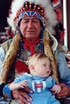 A Native American woman wearing traditional headdress and clothing, holding a young child who is sitting on her lap.