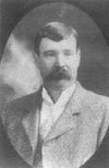 Black-and-white photo of a man with dark hair and a mustache, wearing a suit jacket and shirt, posing against a blurred background.