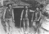 Three men in old-fashioned clothing standing outdoors near a rocky cliff or cave entrance.