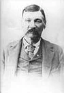 Black and white photo of a man with a beard and mustache, wearing a suit and tie.