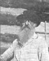 A black and white photo of a man with a beard and glasses, wearing a hat and striped shirt, outdoors with a body of water in the background.
