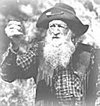 An elderly man with a long white beard, wearing a wide-brimmed hat and plaid clothing, smiling outdoors.