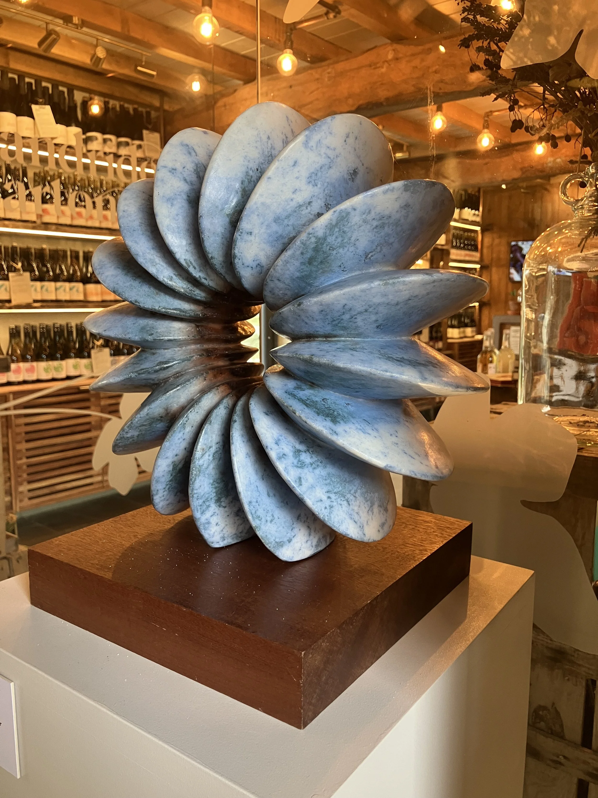 A decorative sculpture made of blue and gray stones resembling shells, displayed on a wooden base in a warmly lit indoor space with shelves of bottles in the background.