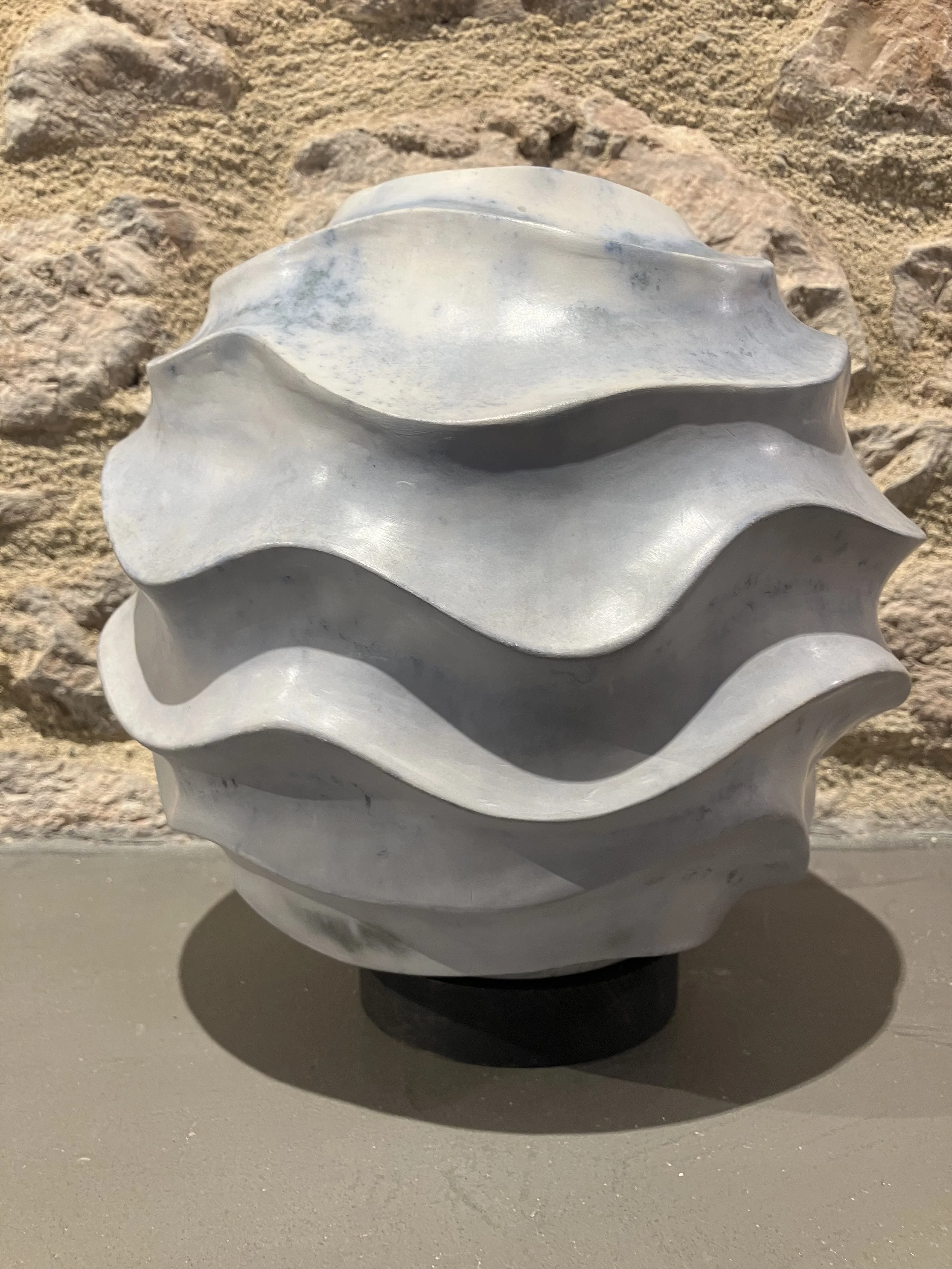 A shell-shaped sculpture or object with a glossy white surface and a sculpted textural design is seen against a rocky, sandy background. It is placed on a flat surface with a shadow underneath.