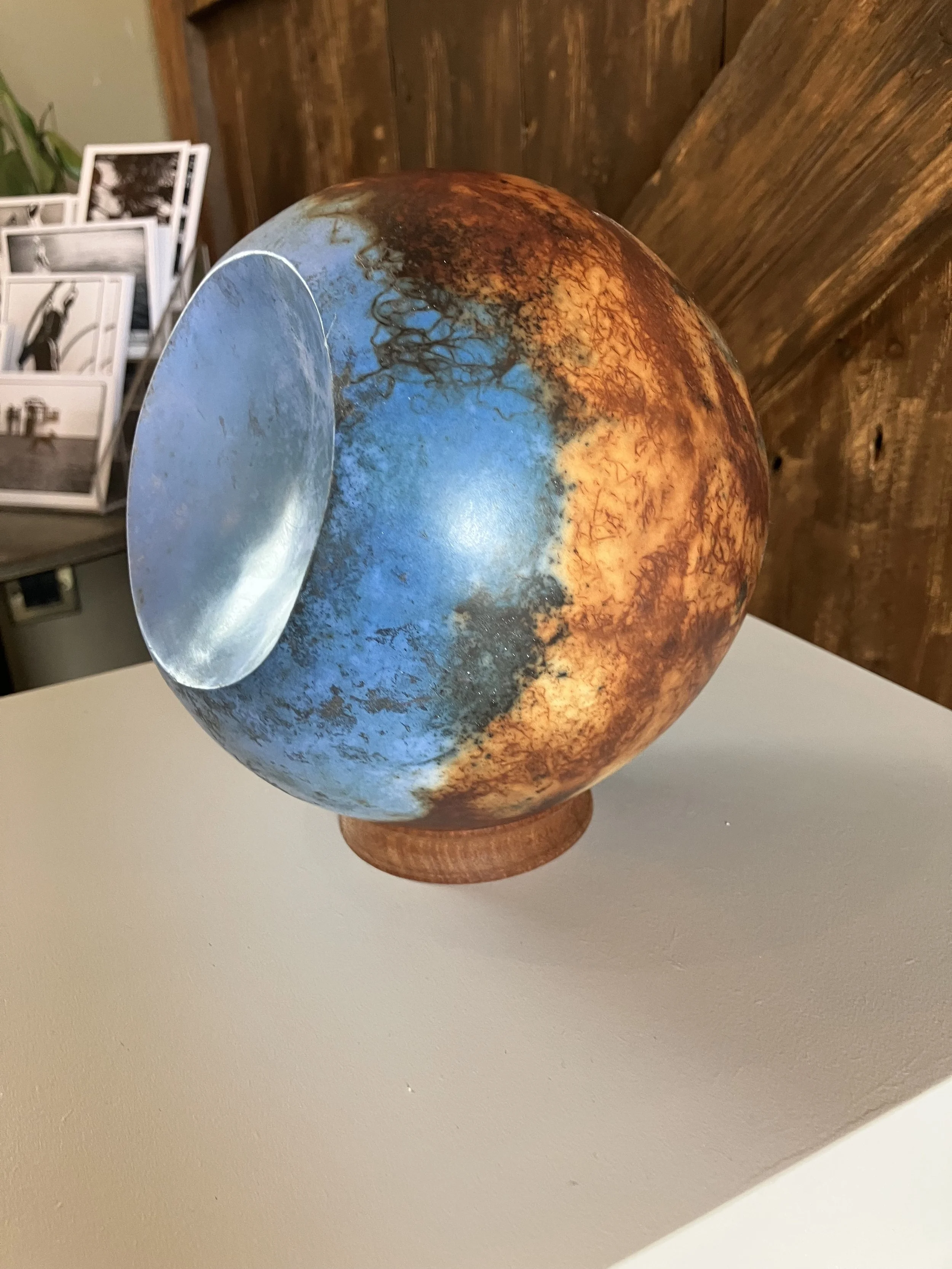 Decorative spherical object with a mix of vibrant blue and warm orange colors, resting on a small wooden base, placed on a white surface with a background of brown wooden wall and photographs on a white photo stand.
