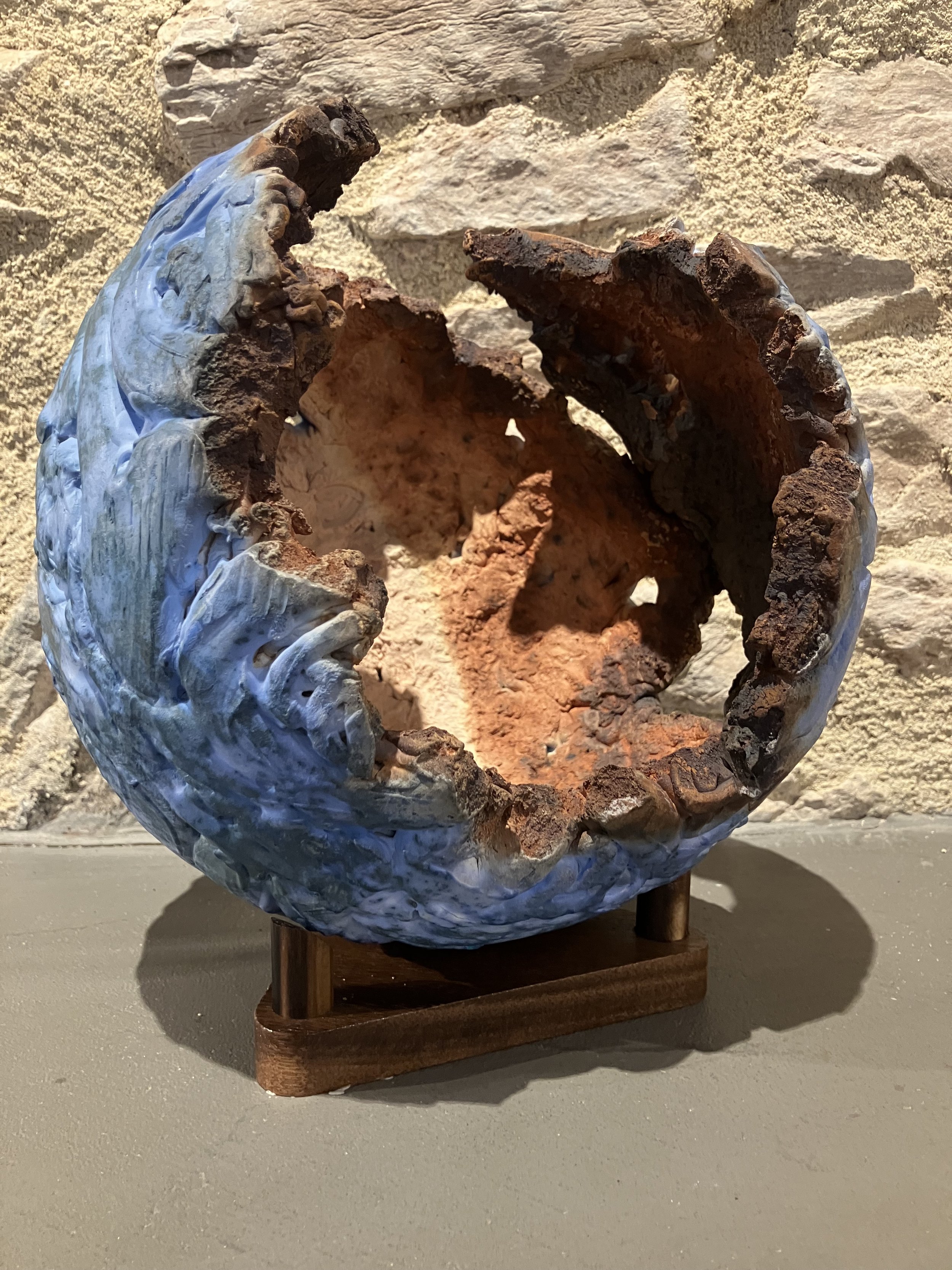 A sculpture of a large oyster with a pearl inside, made of mixed materials including copper and possibly plastic, mounted on a wooden stand, against a rough, beige stone wall.