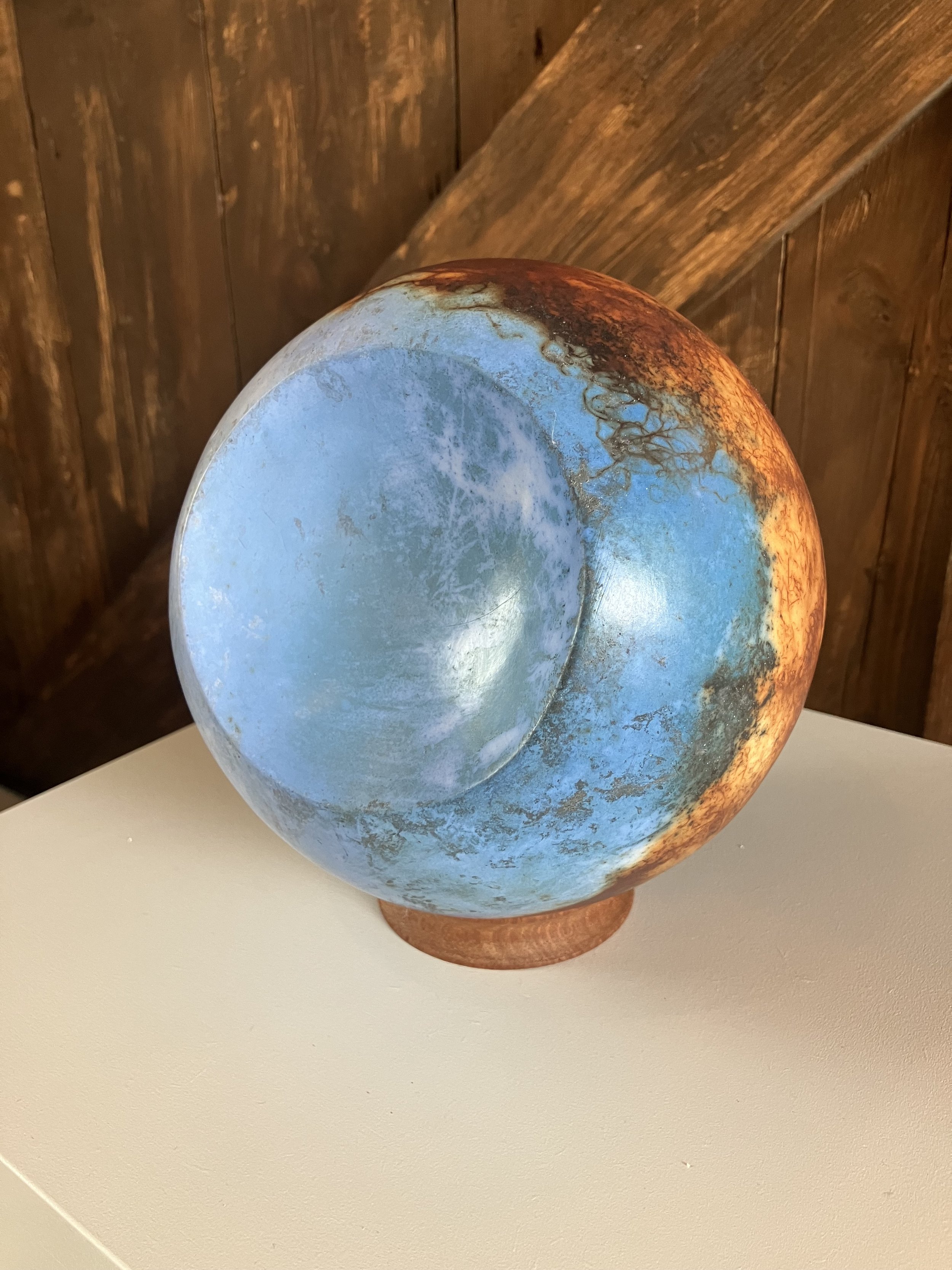 A decorative vessel with a ceramic or pottery finish, featuring a gradient of blue, orange, and brown colors, placed on a white surface against a background of wooden wall panels.