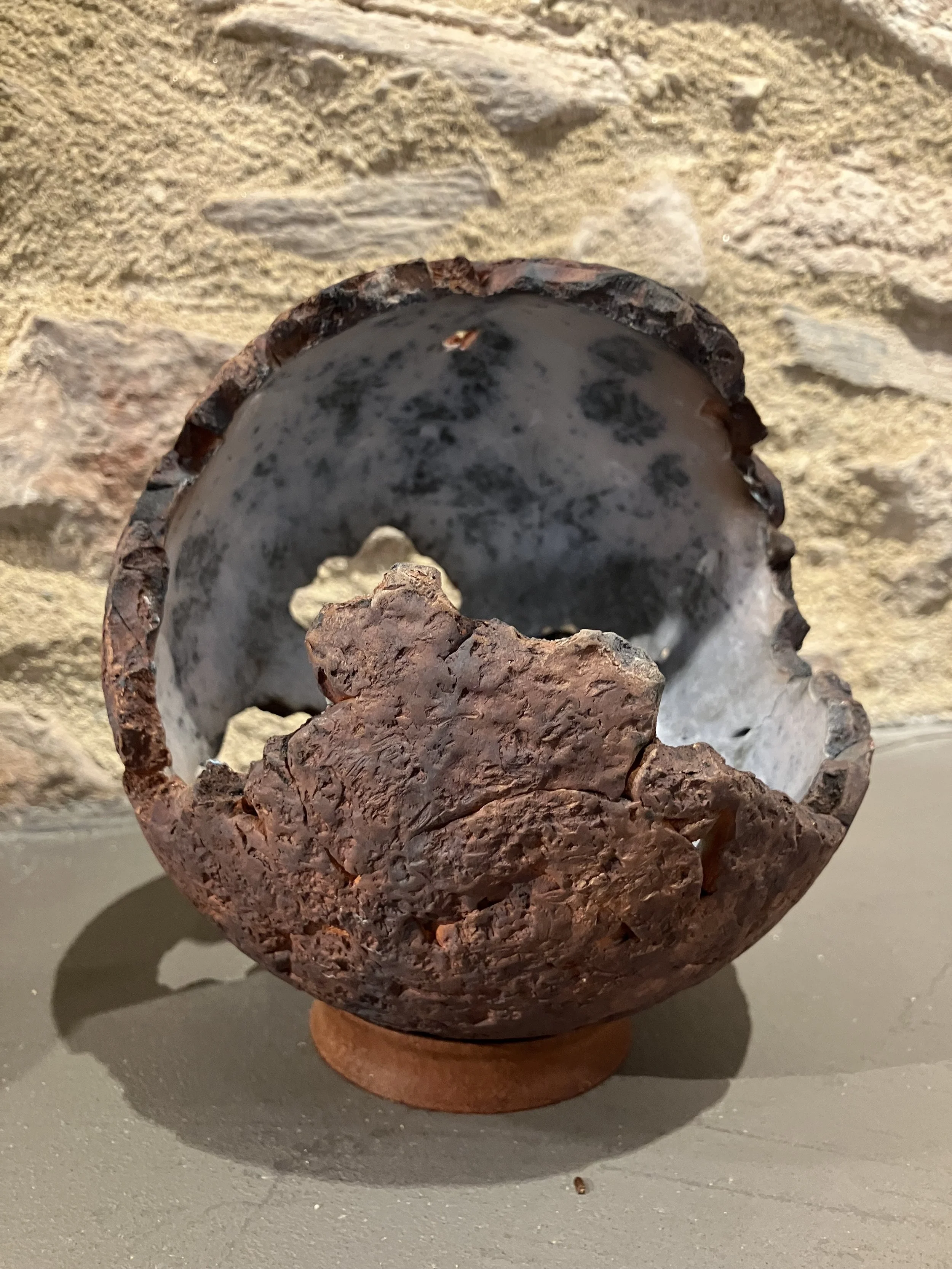 A round ceramic or clay vessel with a rough, reddish-brown, coral-like exterior, resting on a base against a background of a stone wall.