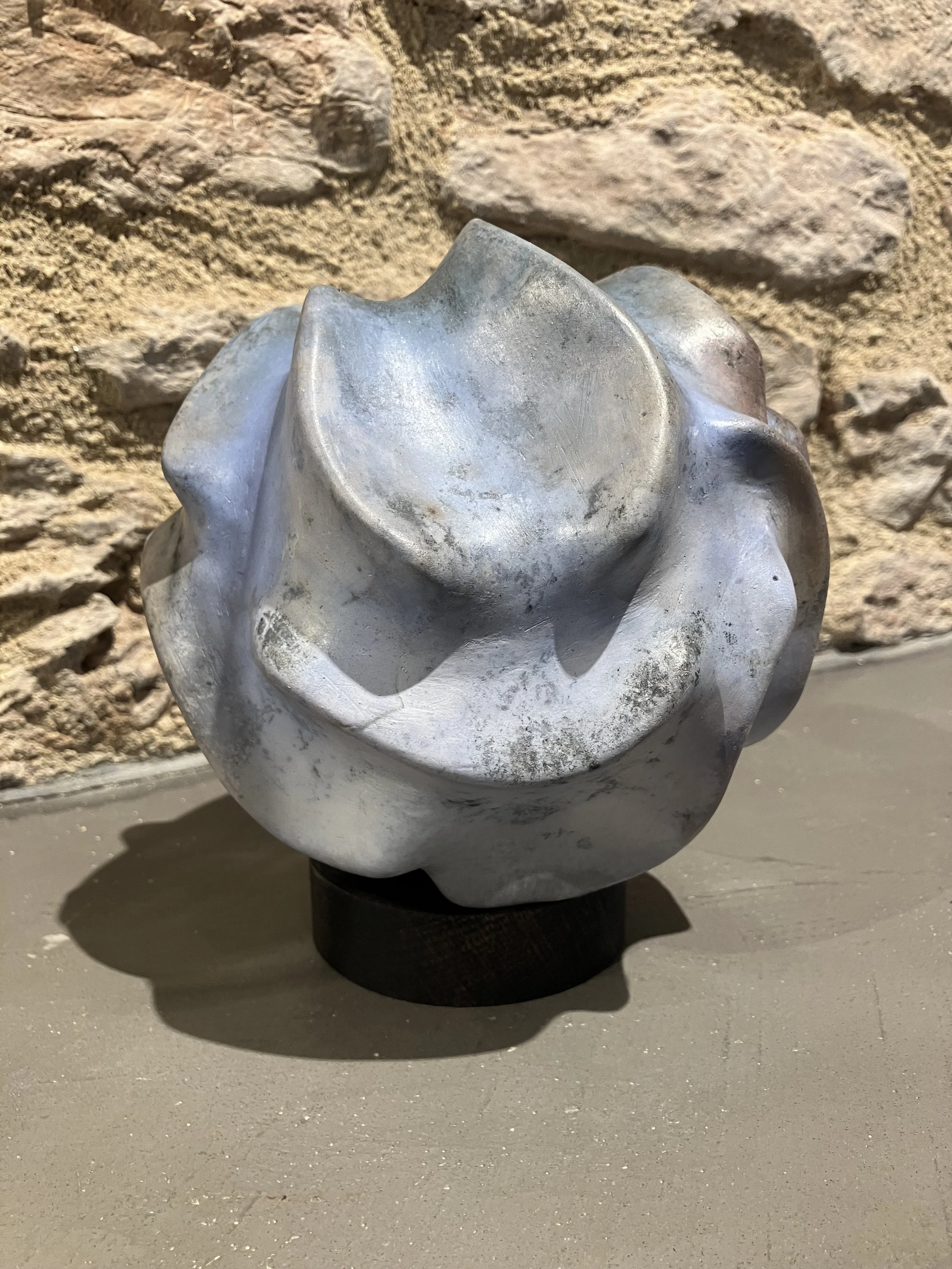 A metallic sculpture resembling a flower or organic shape, placed on a black pedestal against a textured brick wall background.
