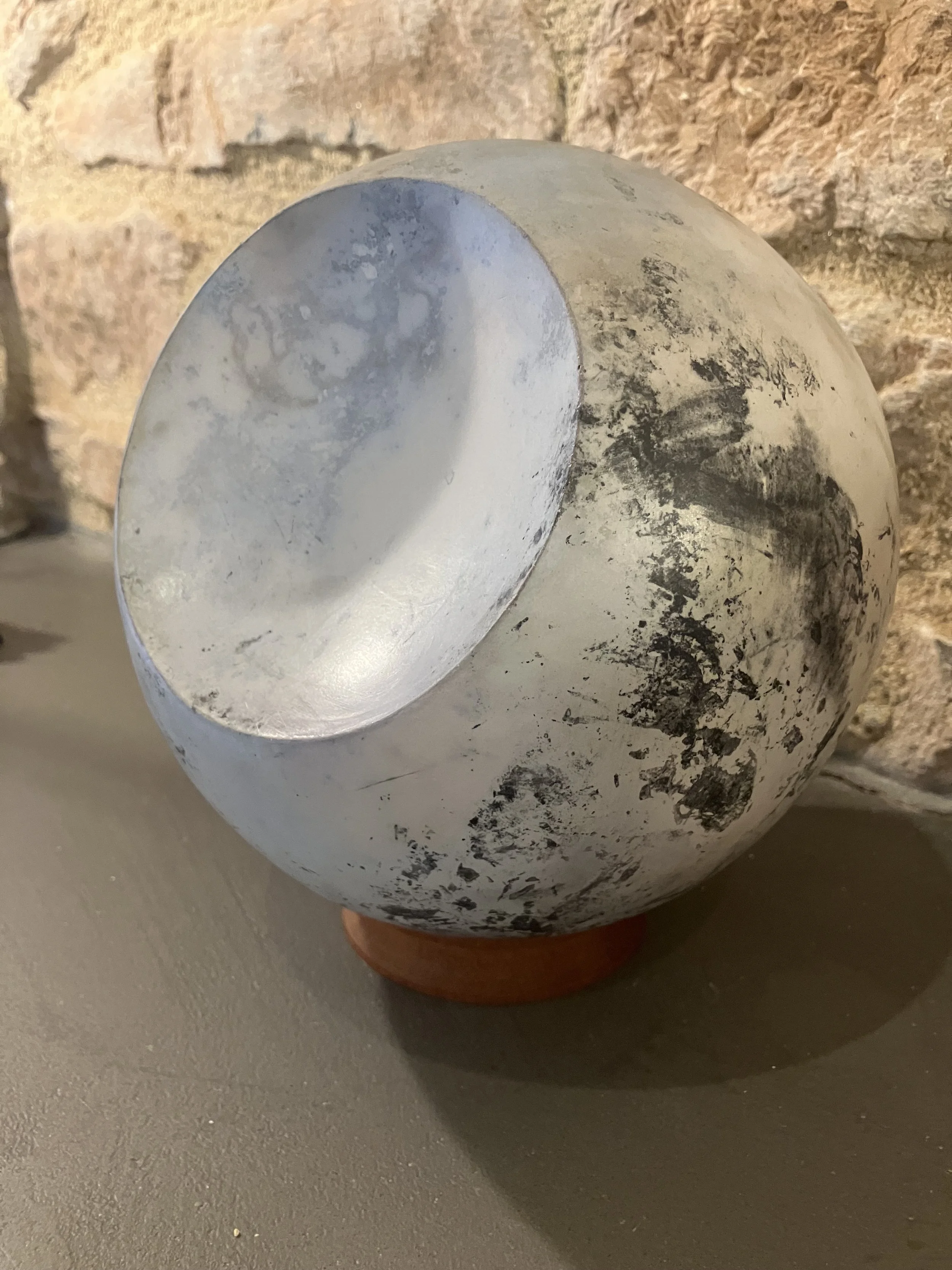 Close-up photo of a partially blackened white spherical object with a hollowed-out top, resting on a reddish-brown base and positioned on a light gray surface with a textured brick wall background.