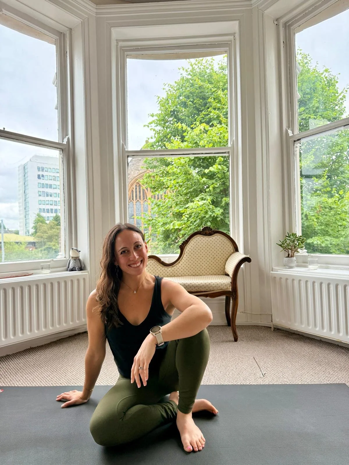A woman practicing yoga on a blue mat in a sunlit room with large windows and green potted plants.