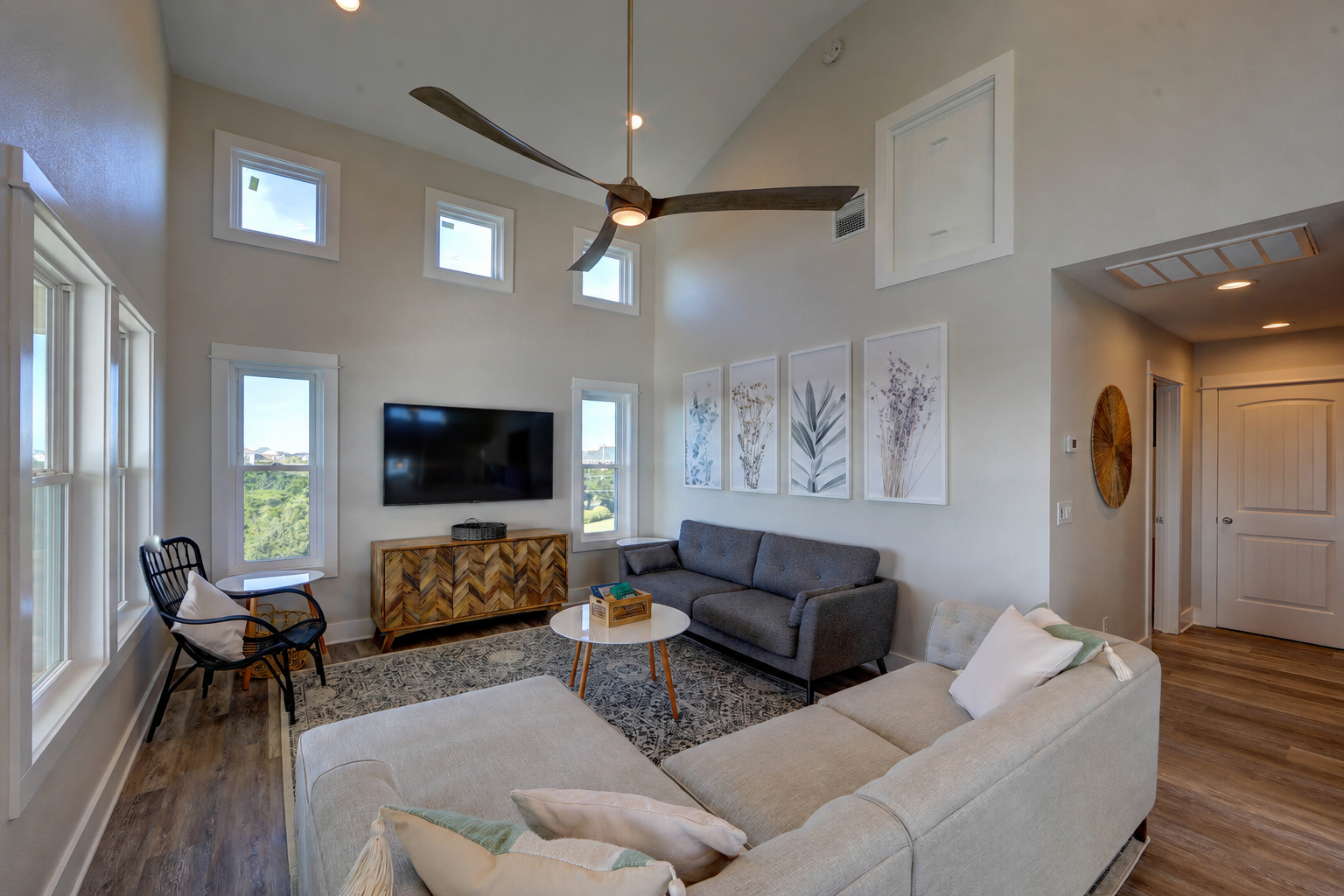 Living room with a beige sectional sofa, a gray sofa, and a black chair with a white cushion, all on a patterned rug. Four framed botanical prints hang on the wall, a wooden sideboard, a wall-mounted flat-screen TV, and a ceiling fan.