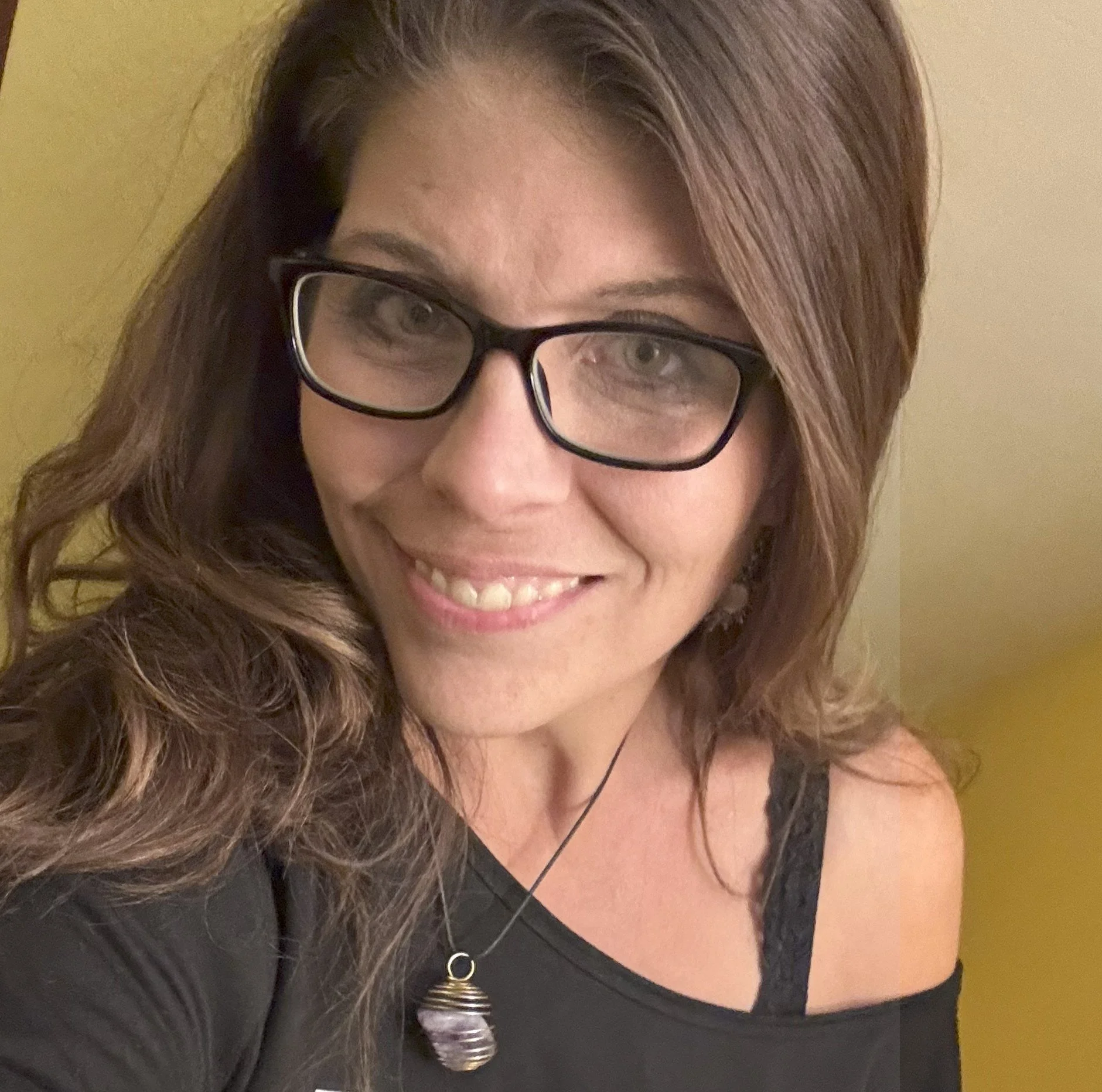 Smiling woman with glasses and wavy brown hair, wearing a black top with a lace strap and a necklace with a purple pendant, standing against a yellow wall.