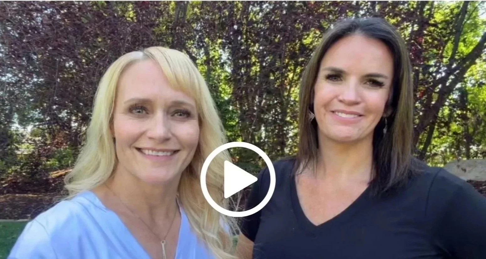 Two women smiling outdoors with trees and greenery in the background.