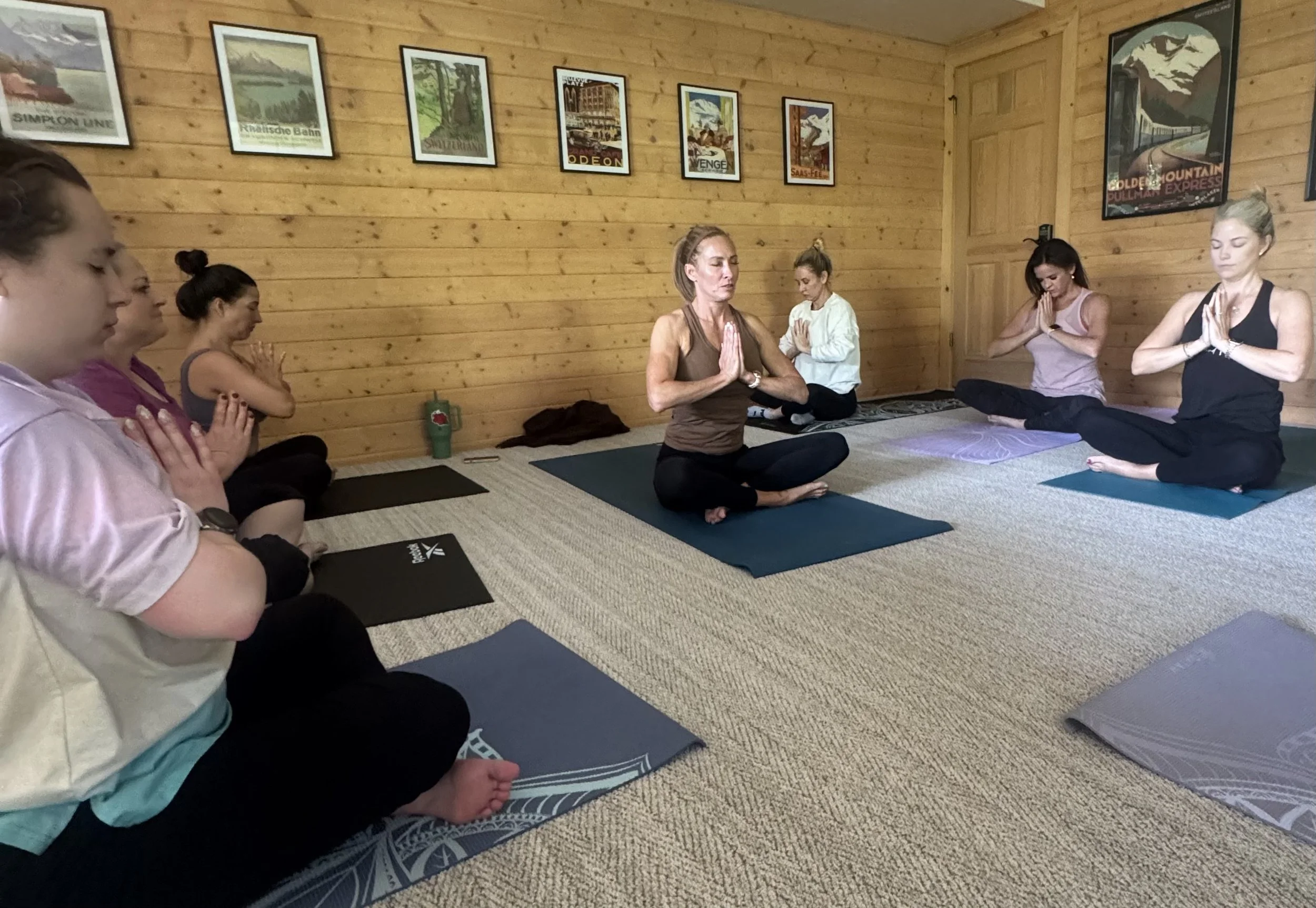 Group of women practicing yoga in a wooden-paneled room, sitting cross-legged with hands in prayer position, eyes closed or looking down, on yoga mats.