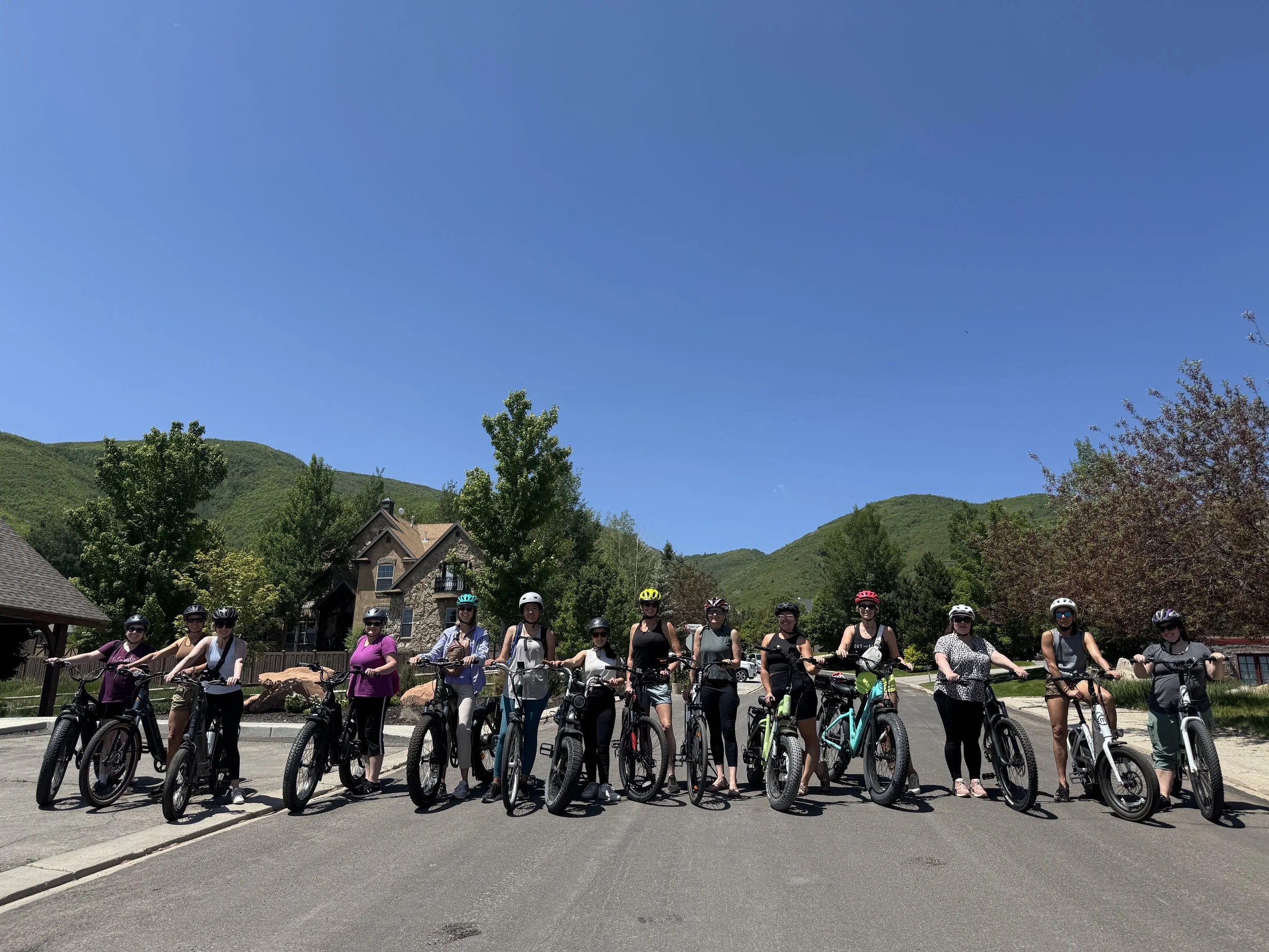 A group of women standing with their bikes on a paved road in a scenic mountainous area on a sunny day.
