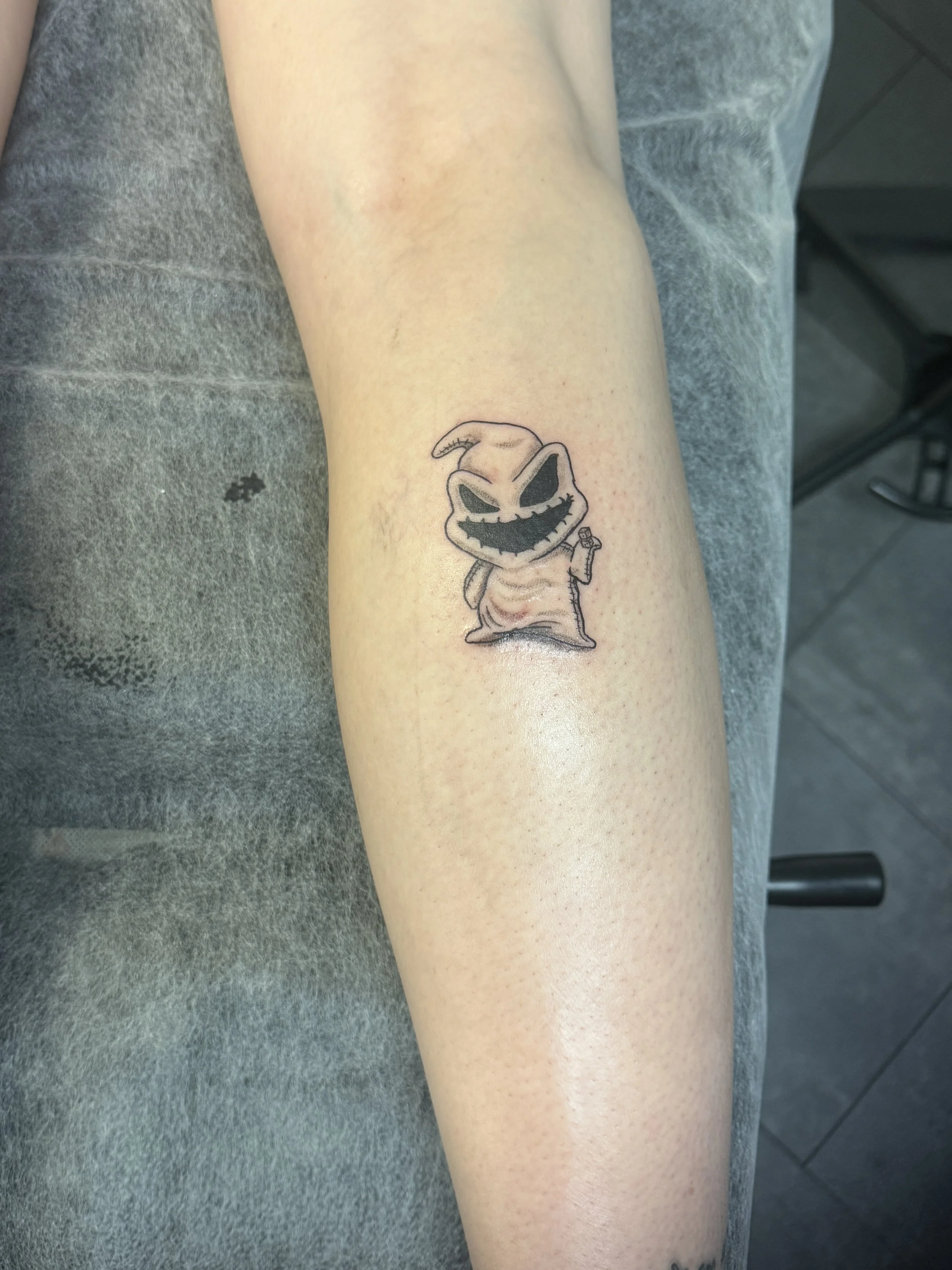 Tattoo of a cartoonish, mischievous ghost with a large, wide grin, sharp teeth, and dark eyes, holding a small item in one hand, on a person's forearm.
