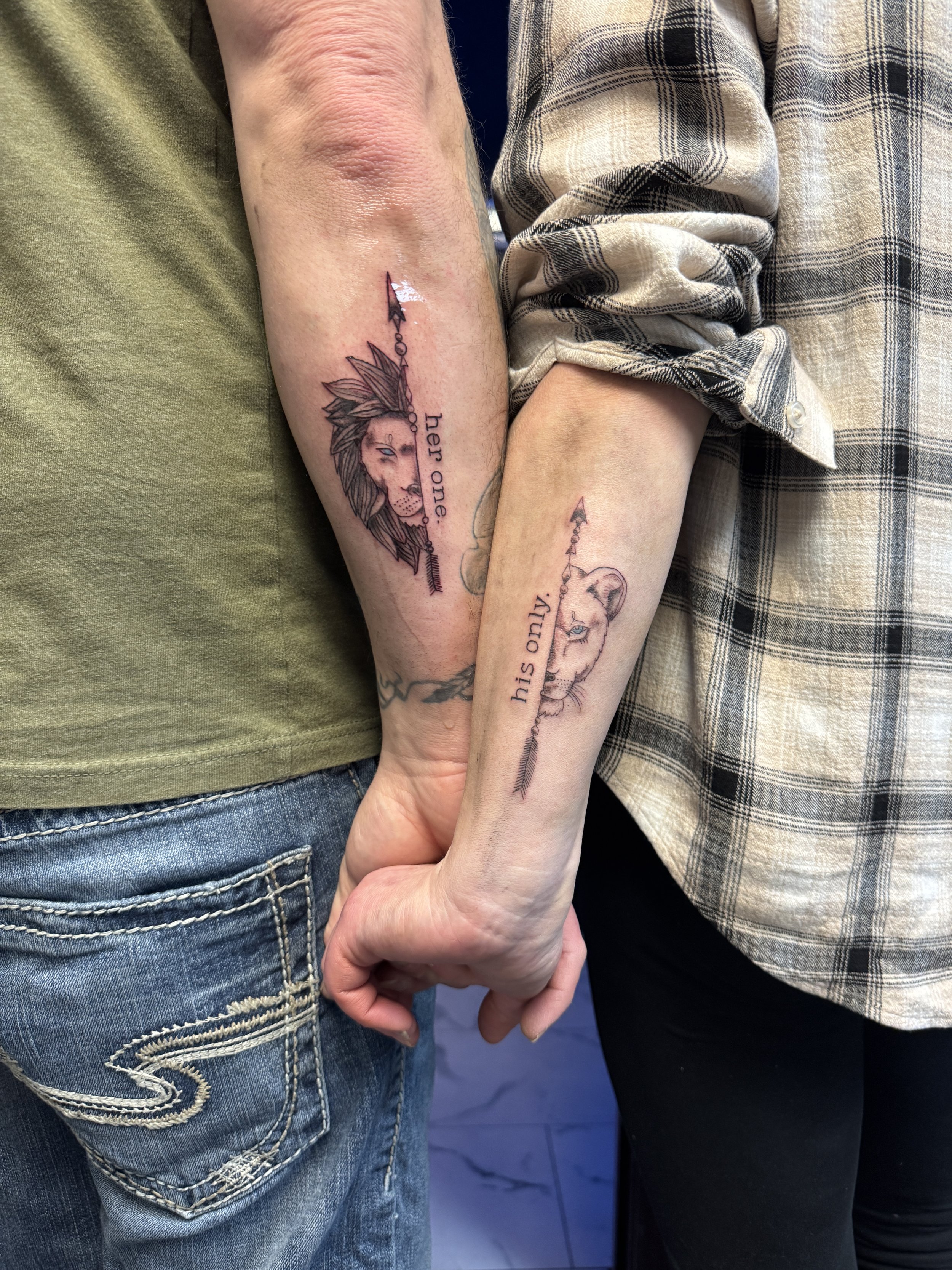 Tattoos of a lion and a lioness, each with an arrow through their heads, with the words "her one" and "his one" beside each tattoo, on the arms of two people holding hands.