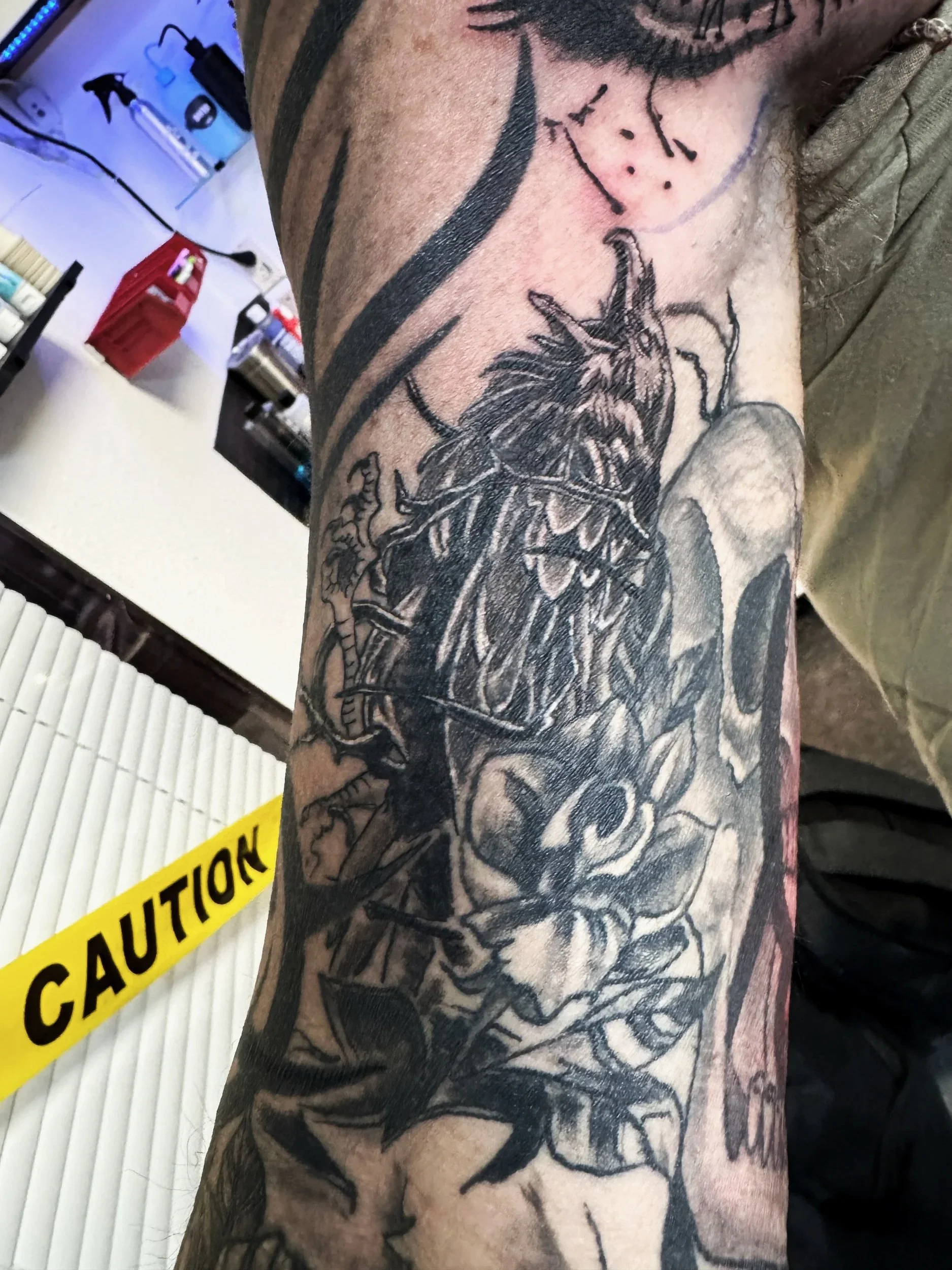 A person's arm with an intricate black and gray tattoo of a dragon, with a caution tape in the background reading 'CAUTION' and various objects on a white surface, including a red box and a blue light.
