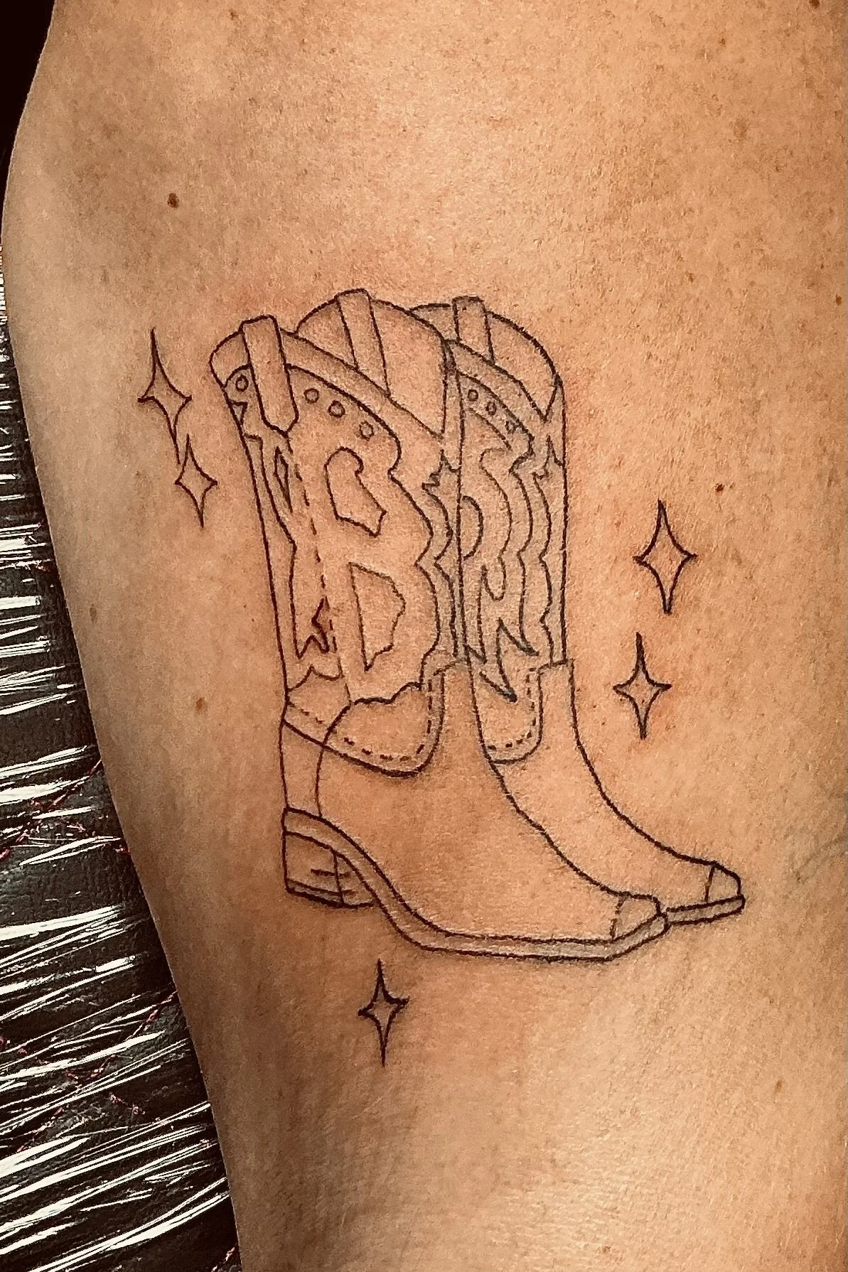 Tattoo of a cowboy boot with a large guitar pick, outlined with minimal detail, surrounded by small star-shaped sparkle symbols.