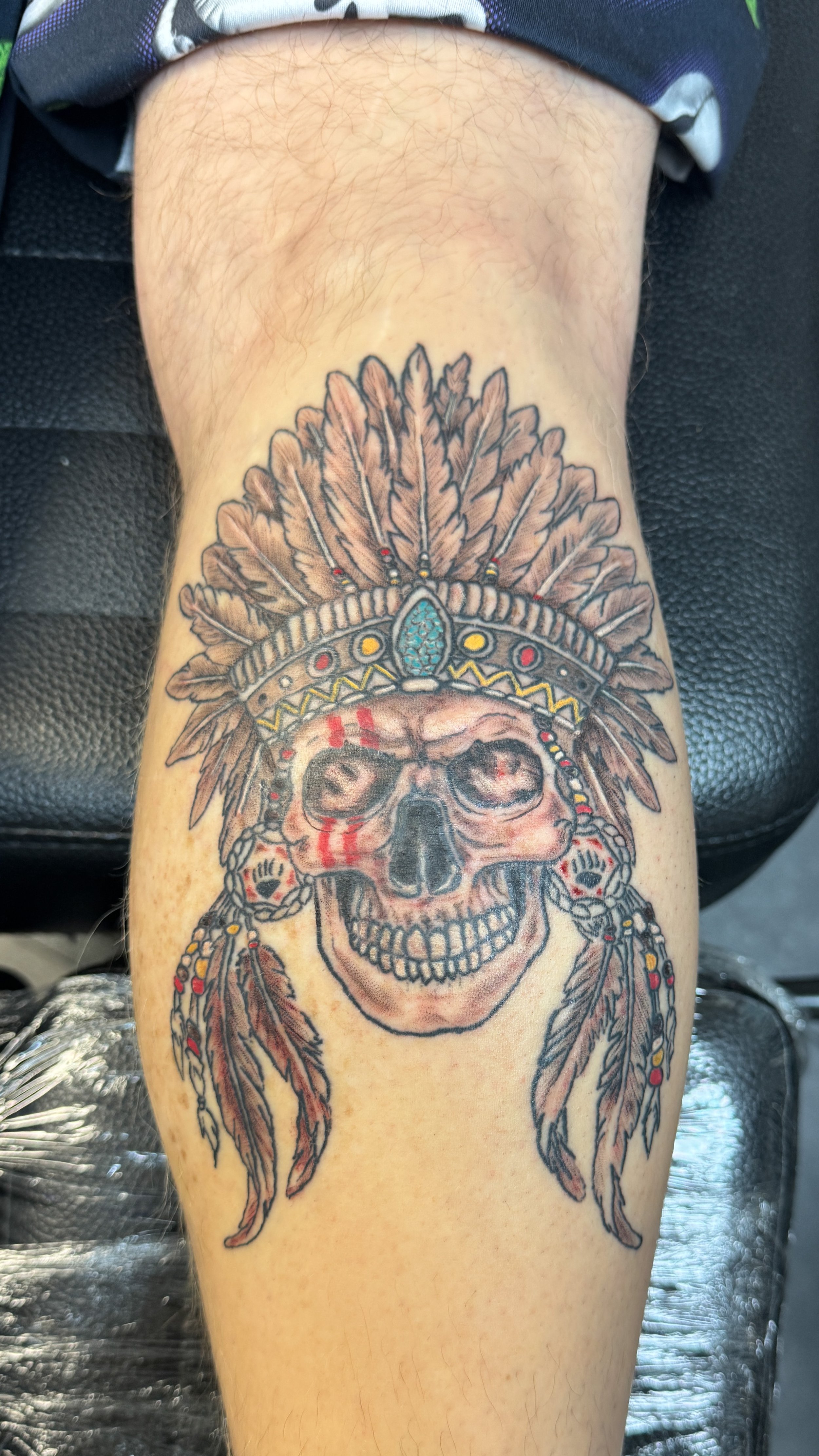 Tattoo of a skull wearing a Native American headdress with feathers, with red markings on the face and beads in the headdress and earrings.
