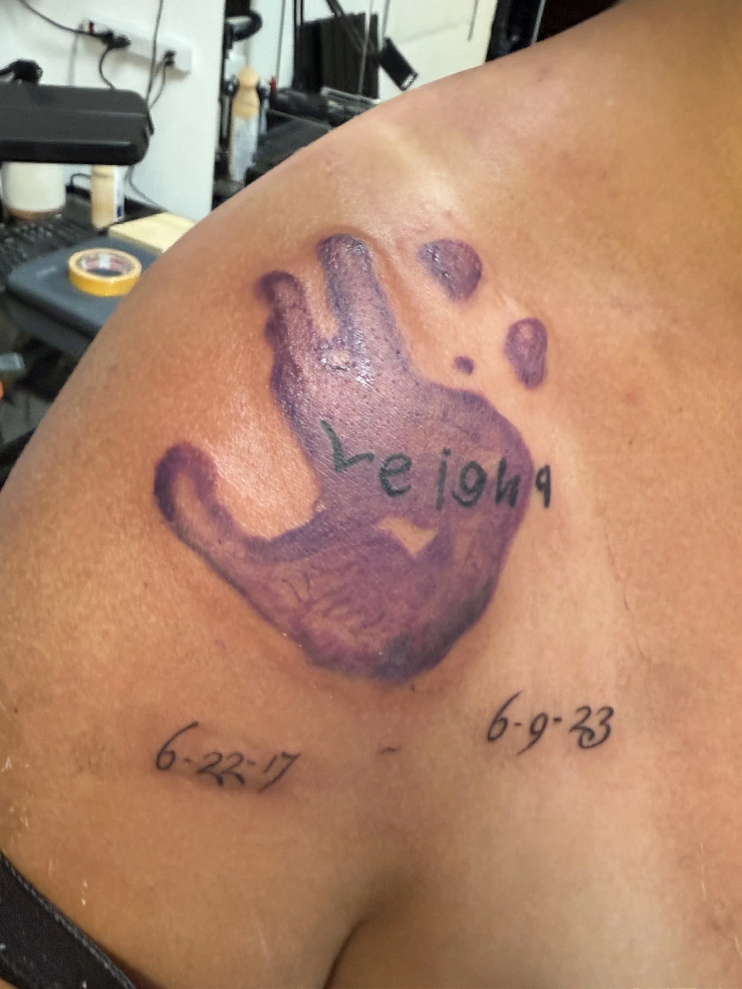 Tattoo of a cat's paw print with the name 'Leiah' inside it and the dates '6-22-17' and '6-9-23' beneath on a person's shoulder.