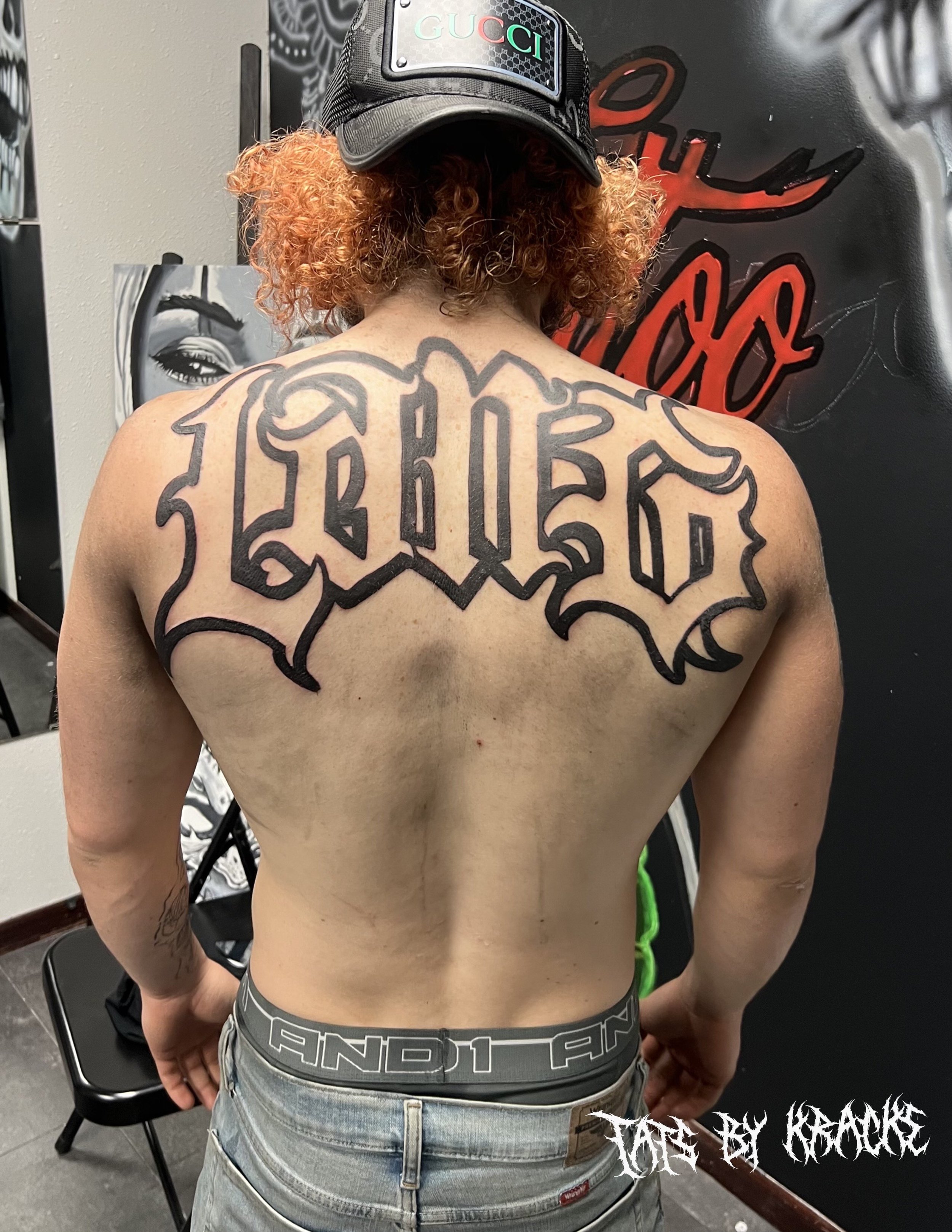 Back view of a shirtless person with large tattoo of the word 'LOYAL' across their upper back, wearing a black Gucci cap, jeans, and standing indoors.
