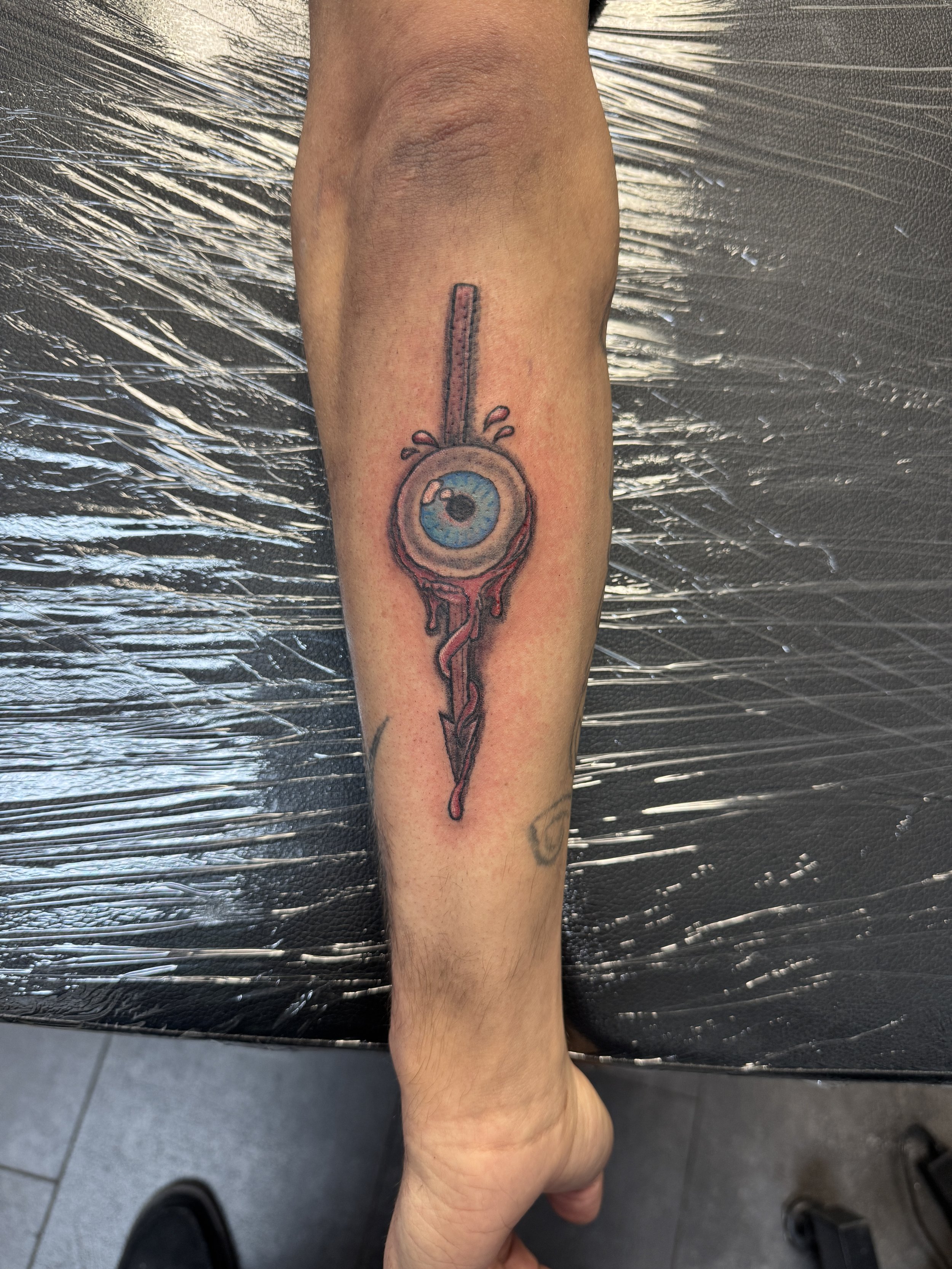 Tattoo of an eyeball with a blue iris and a black pupil, with blood dripping from the bottom, pierced by a vertical knife through the center, on a person's lower leg.