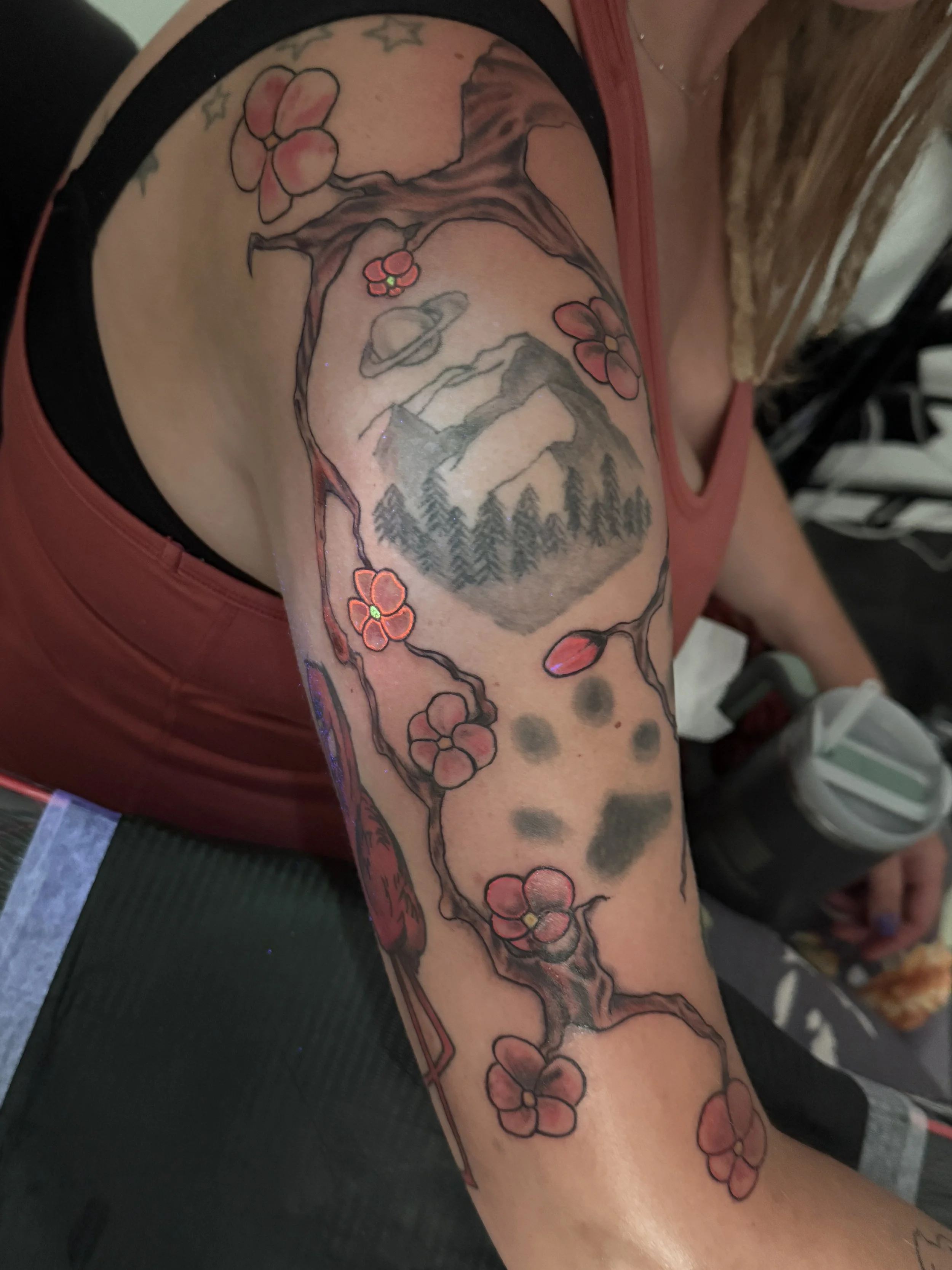 A person with a colorful sleeve tattoo of a mountain landscape, pine trees, a planet with rings, and cherry blossoms on their upper arm.