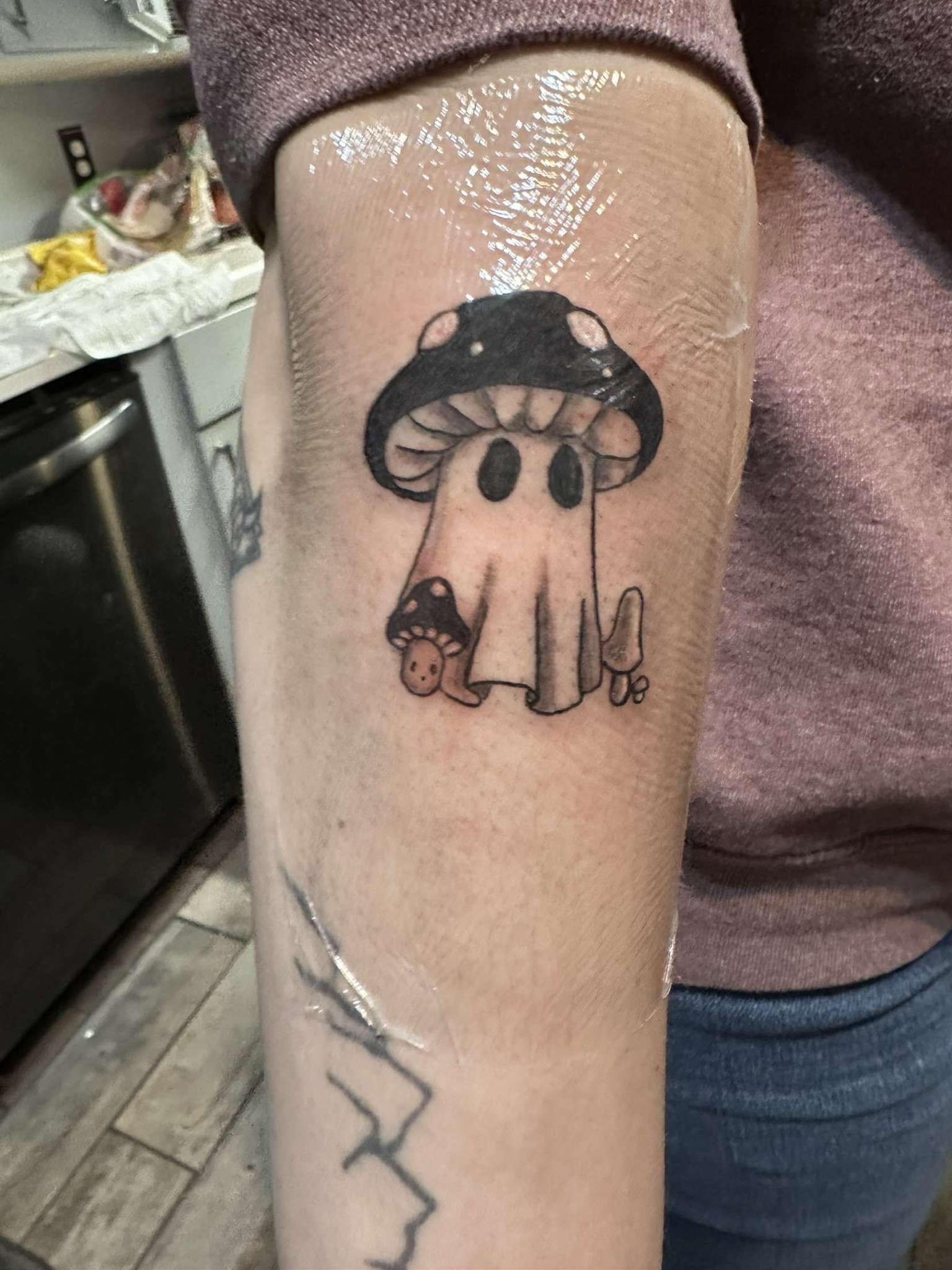 Tattoo of a mushroom with a face, with a smaller mushroom at its base, in black ink on a person's arm.