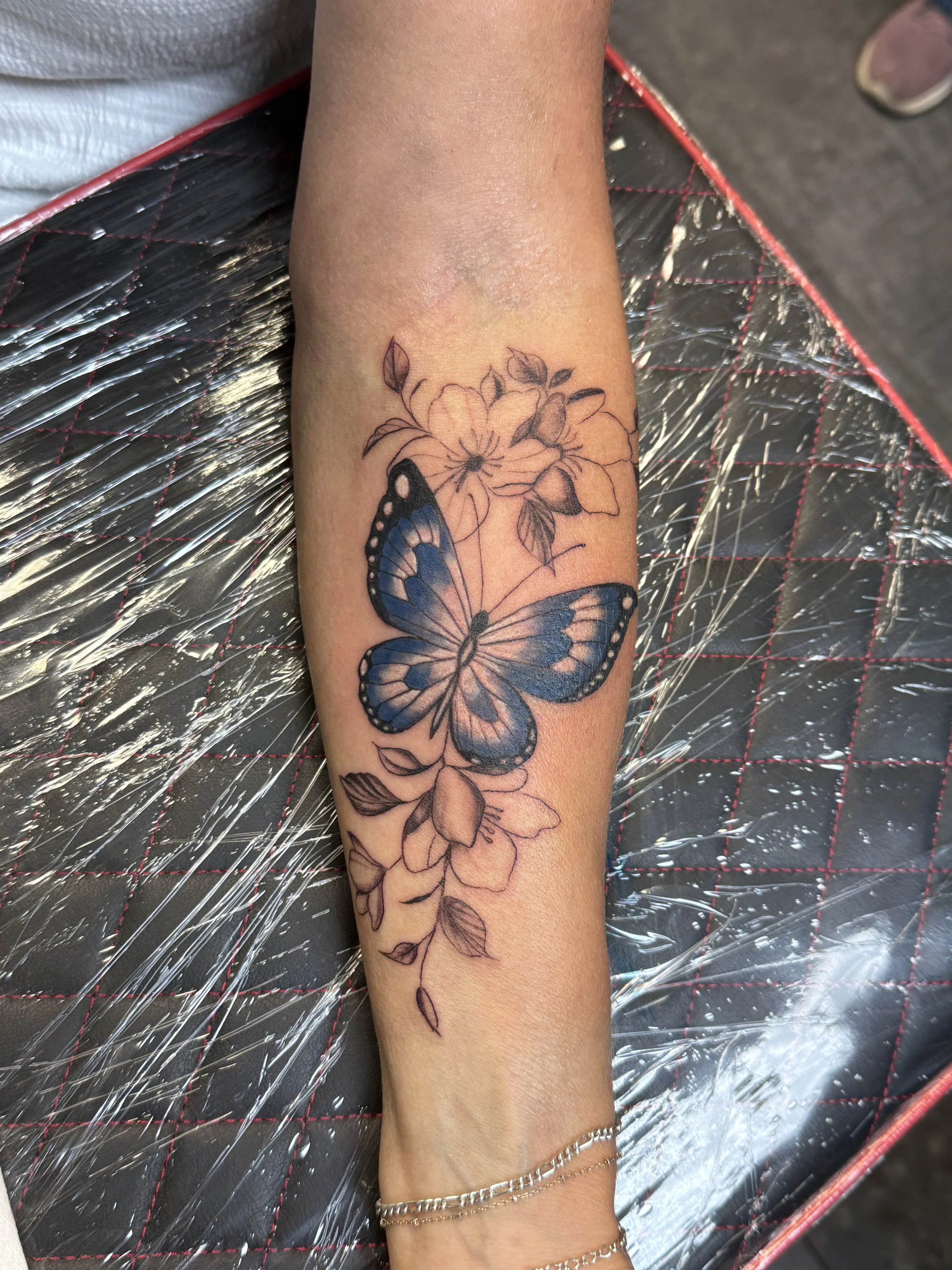 A tattoo of a butterfly perched on a flower with leaves on the forearm.