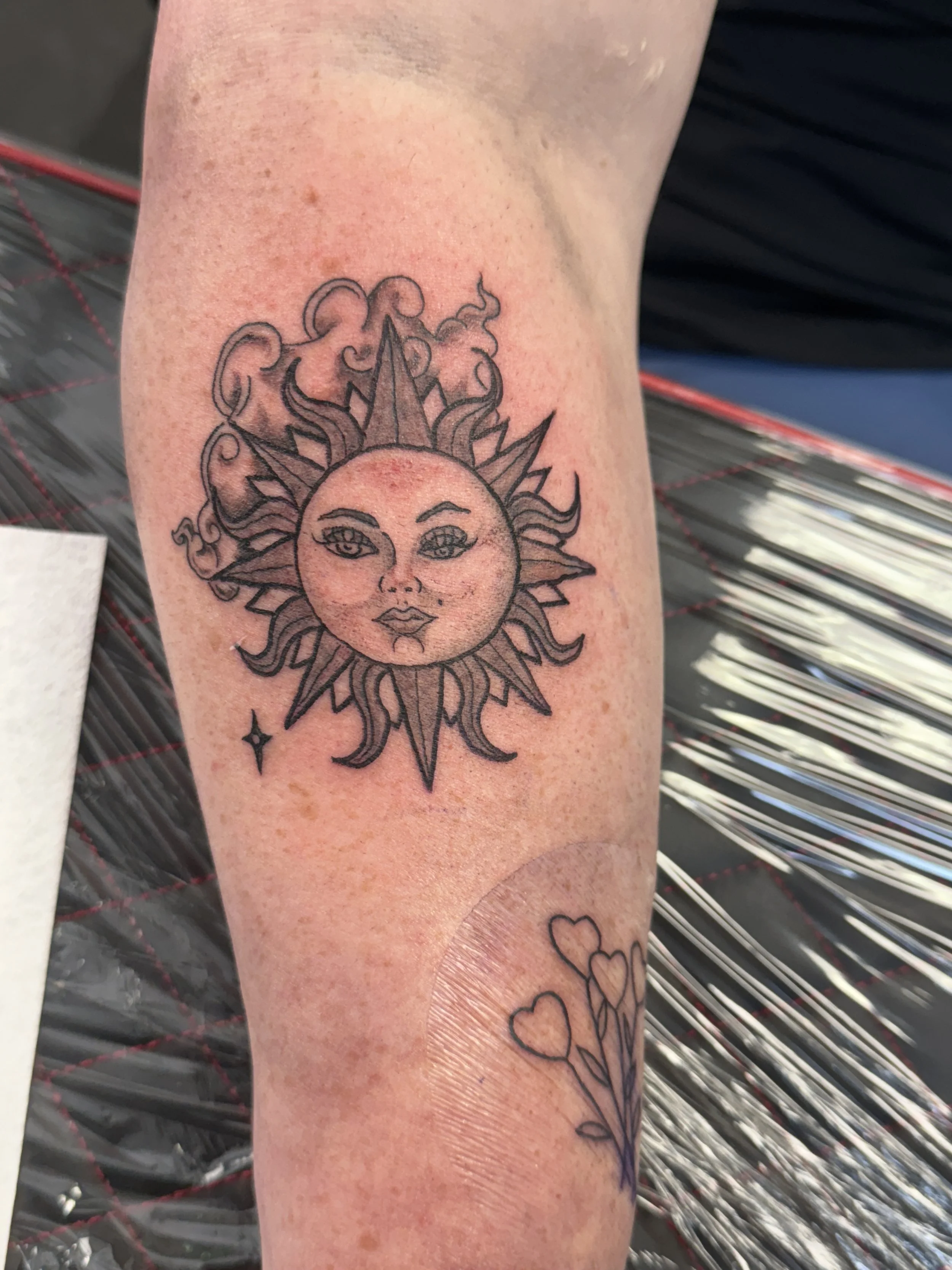 Tattoo of a sun with a face, surrounded by stylized sun rays, on a person's forearm.