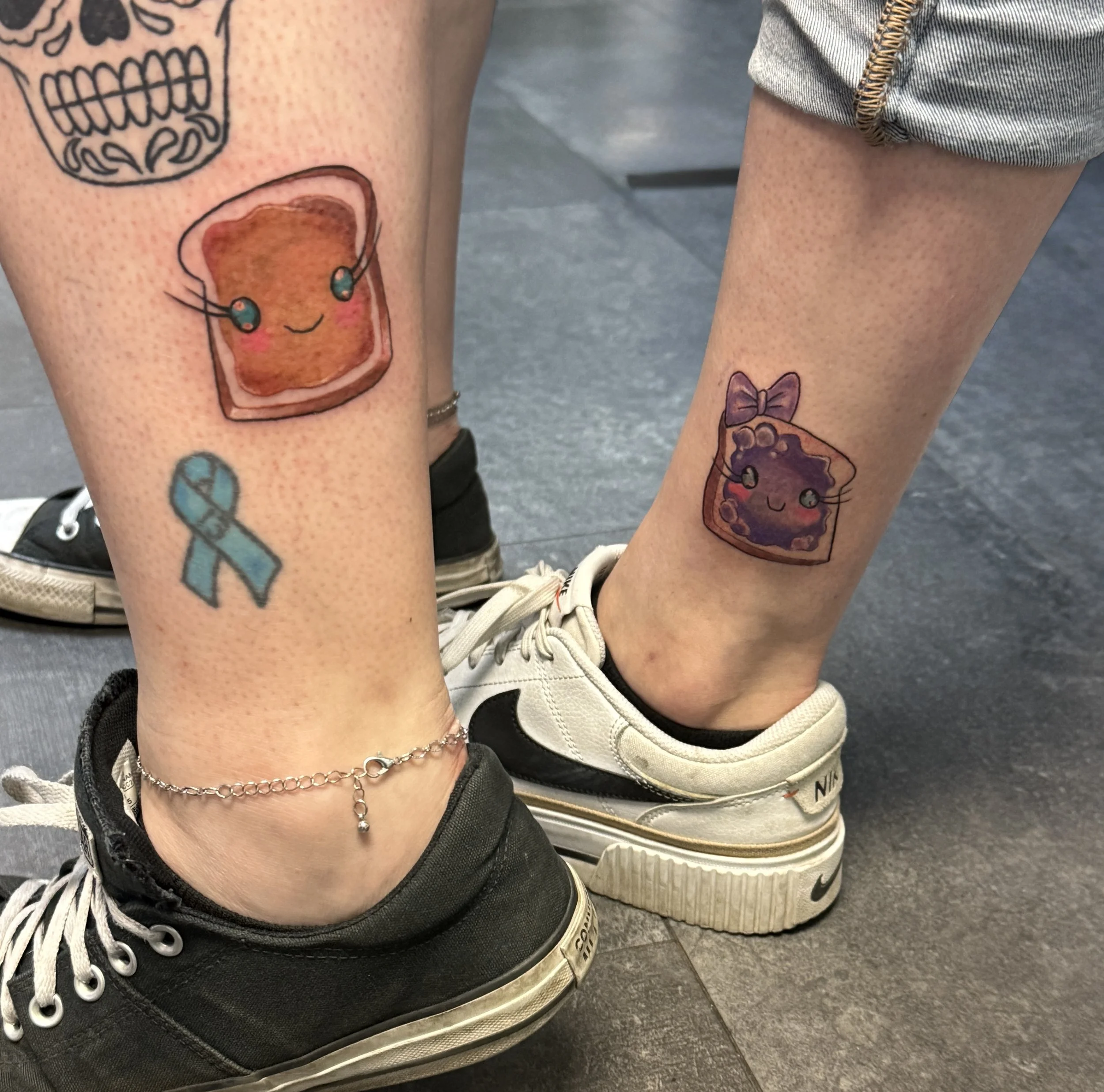 Two legs with colorful tattoos of animated food characters, including a toast with headphones on the left leg and a slice of pie with a bow on the right leg, along with a blue awareness ribbon tattoo.