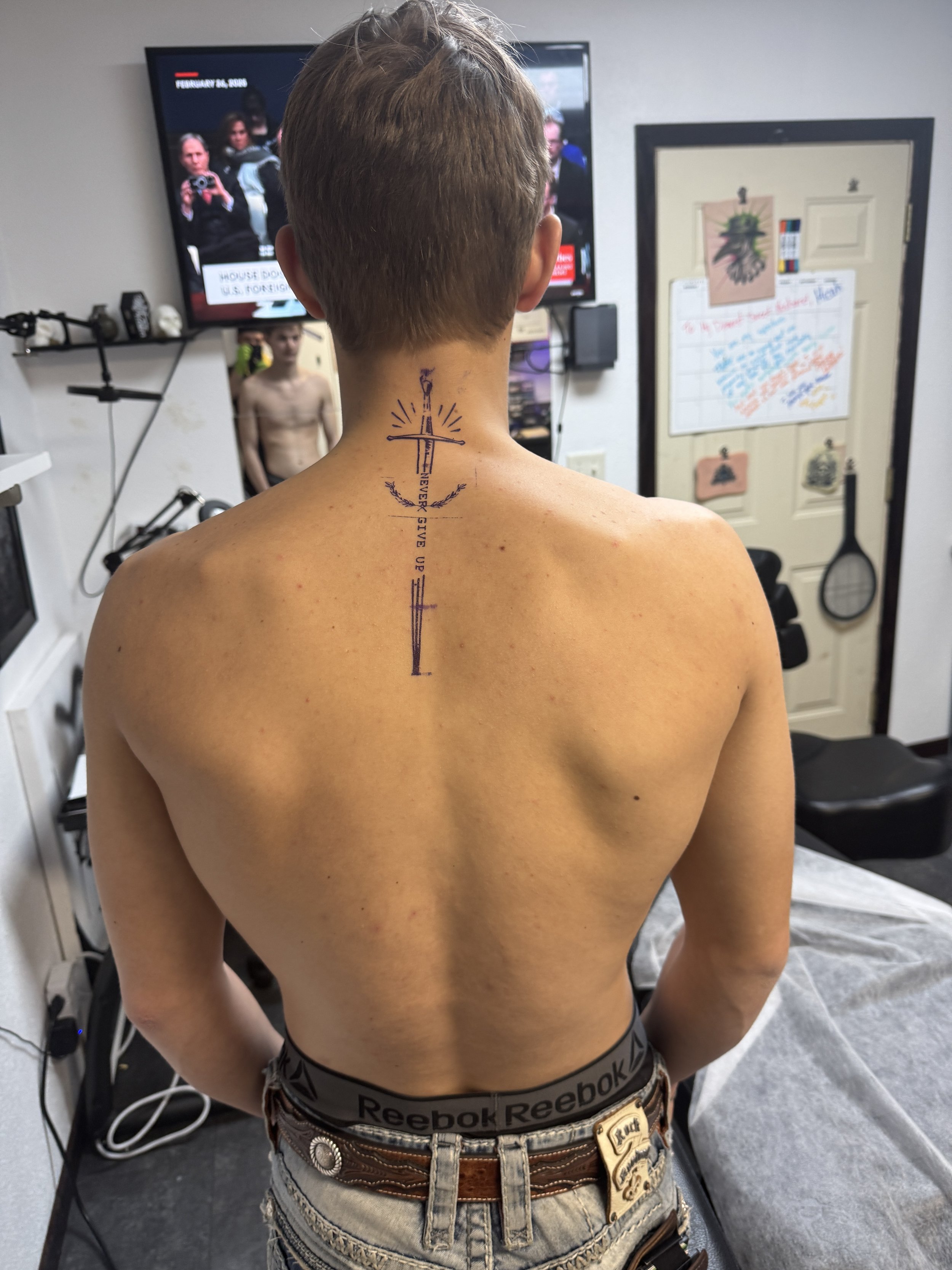 Back of young man with a sword tattoo running down his spine, visible in a tattoo studio with a man in the background and a TV on the wall.