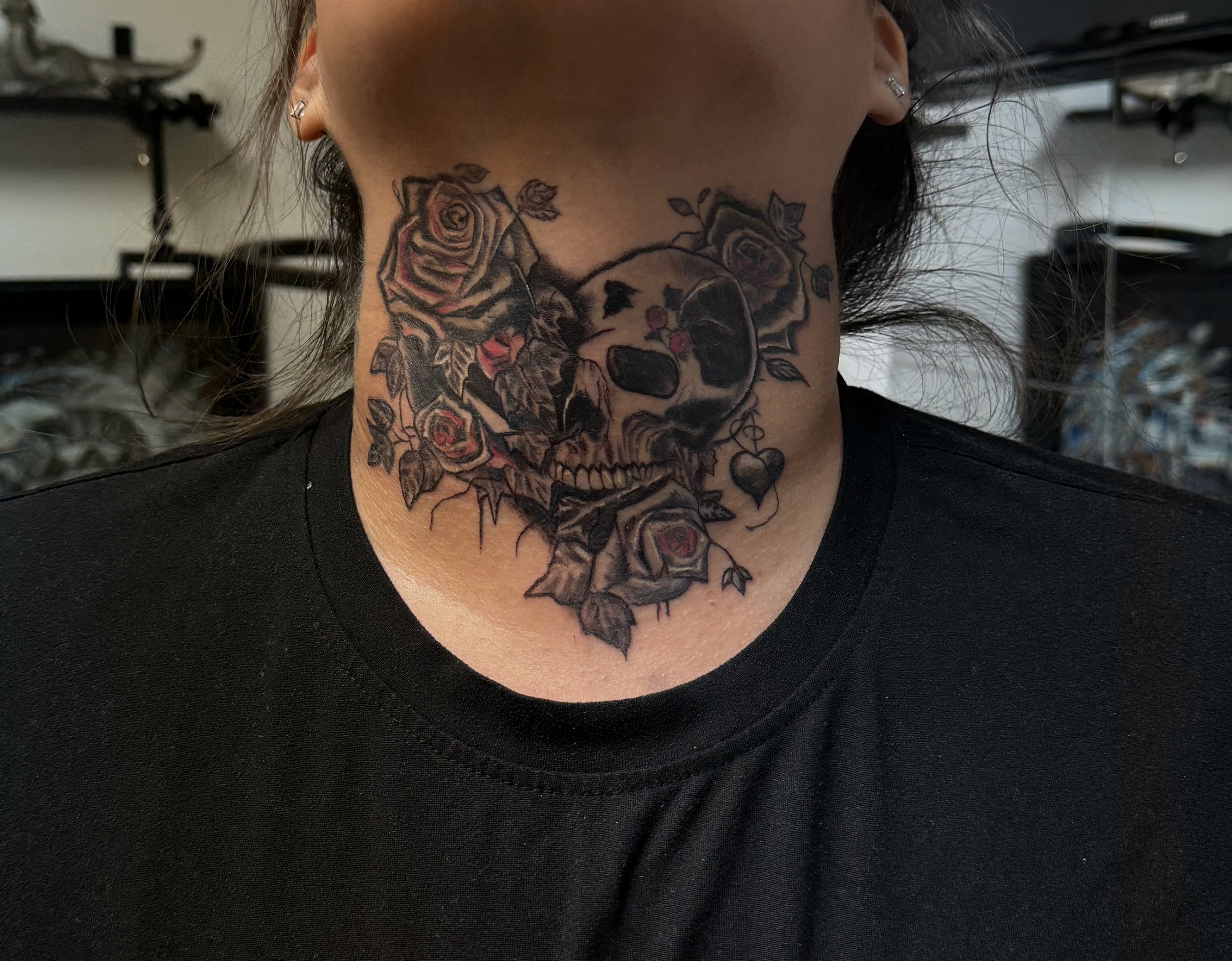Tattoo of a skull surrounded by roses and a heart on a person's neck.