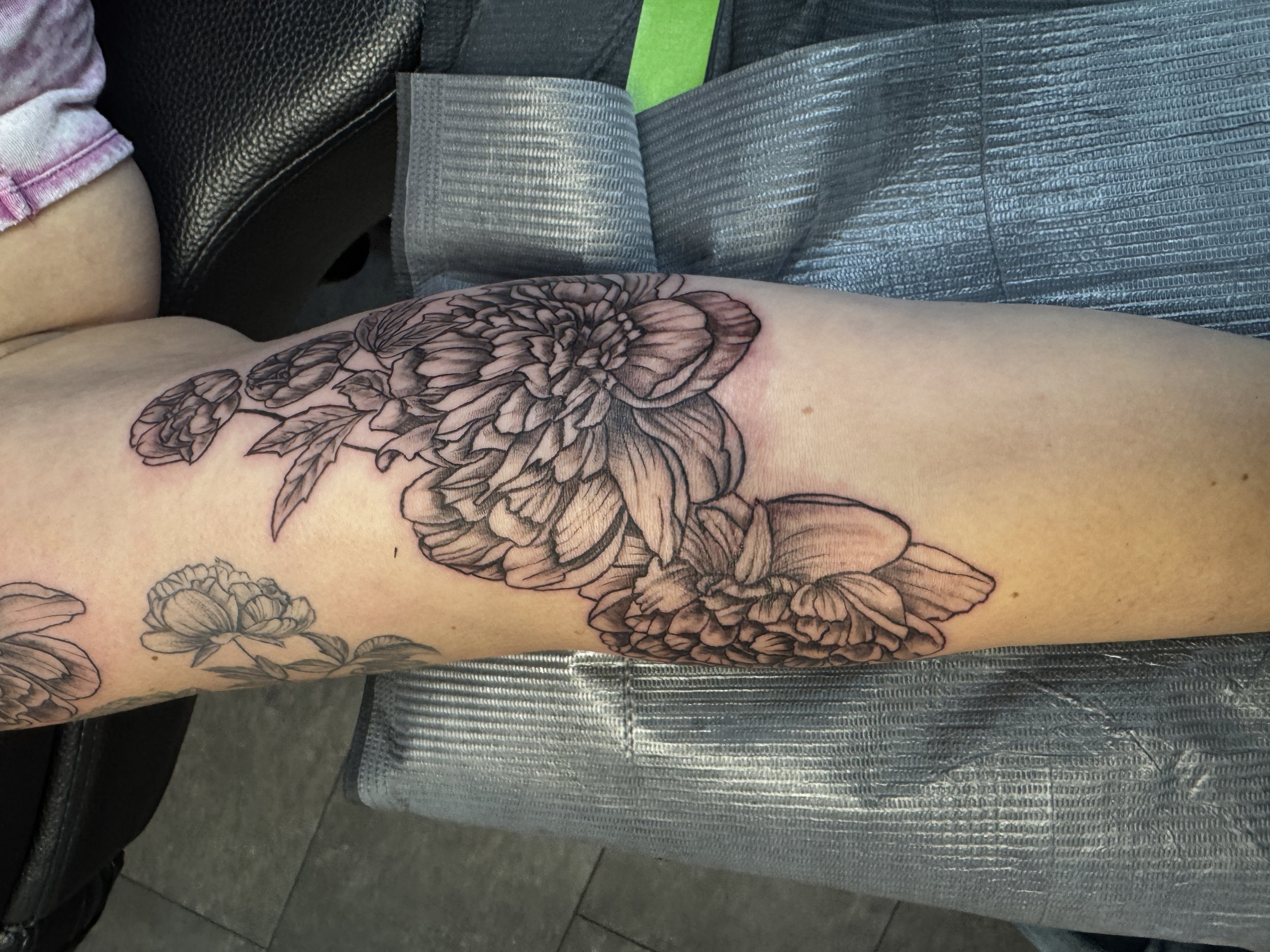 Black and gray floral tattoo on person's thigh, featuring large peony flowers with detailed petals and leaves.